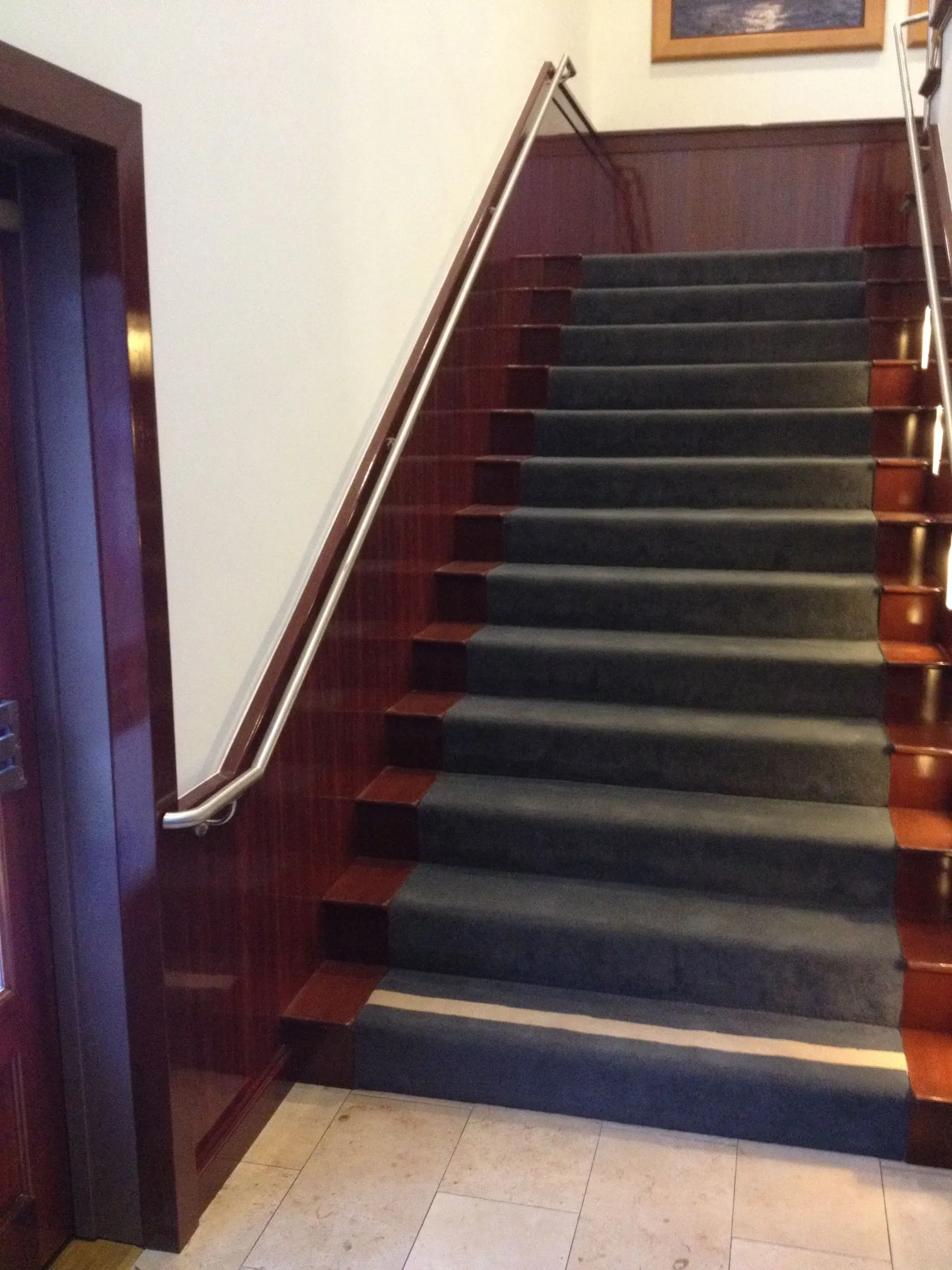 Indoor staircase with dark carpeted steps, wooden banister, and tiled floor at the bottom.