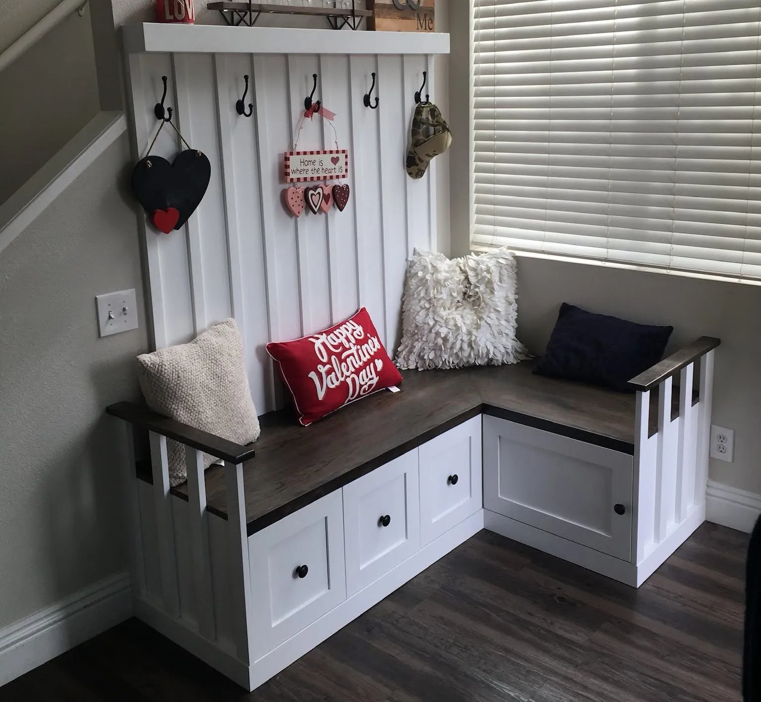 Corner window bench with white storage cabinets, dark wood seat, pillows, and hanging hooks by blinds