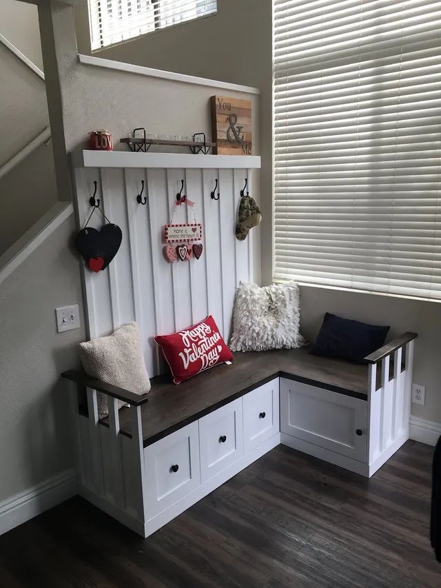 White built-in corner bench with red and patterned pillows, wall shelf, and hanging decor by a window with blinds