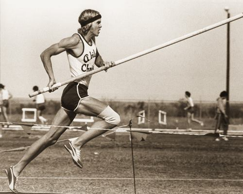 Sepia photo of a pole vaulter sprinting on a track with a pole, preparing for takeoff