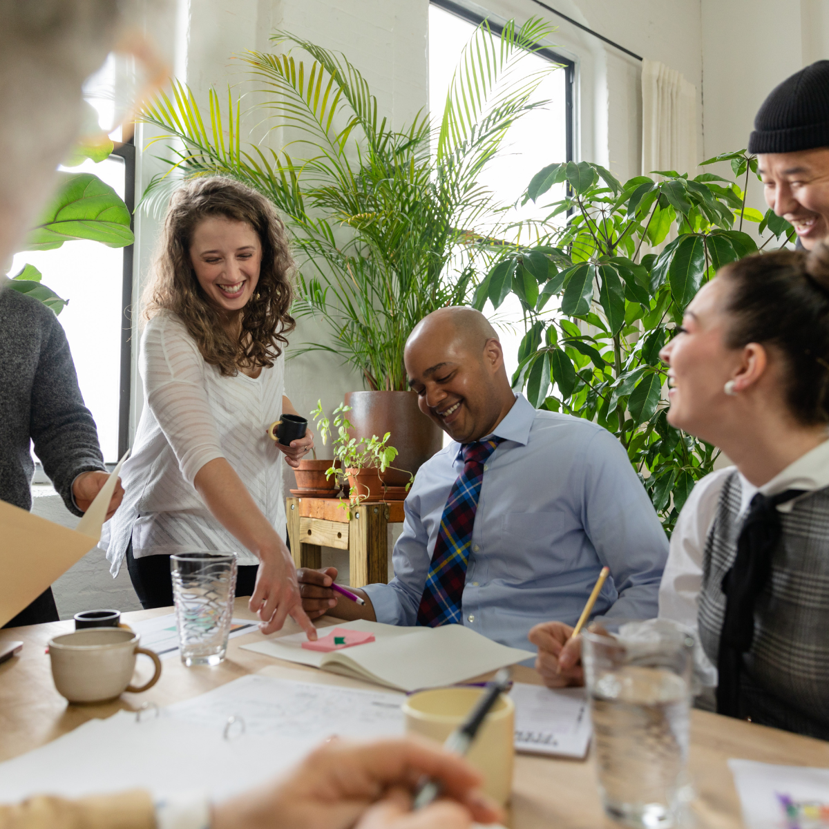 Coworkers in a bright office meeting, smiling and discussing papers around a table with laptops and drinks