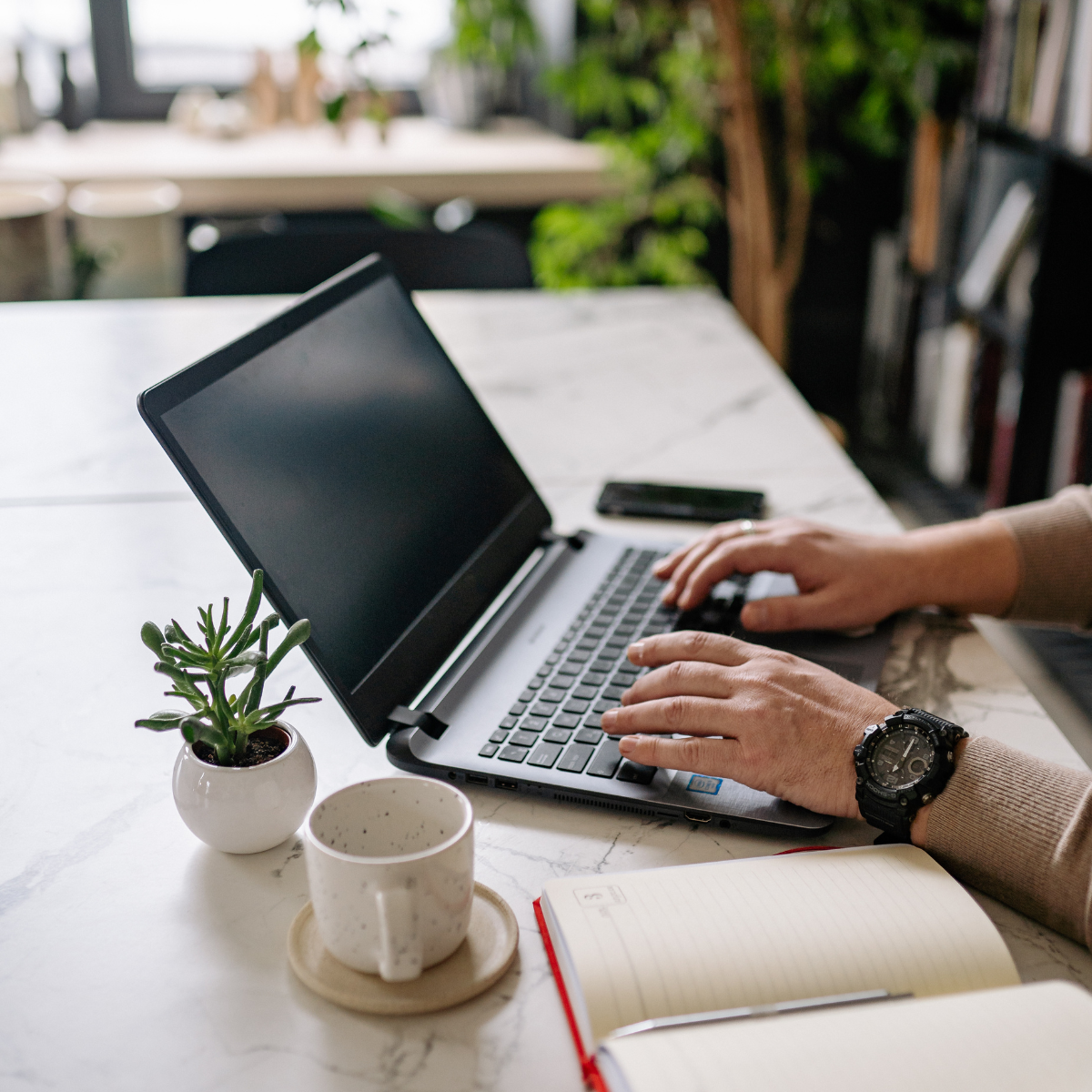 Person typing on a laptop at a desk with a notebook, coffee mug, and small plant.
