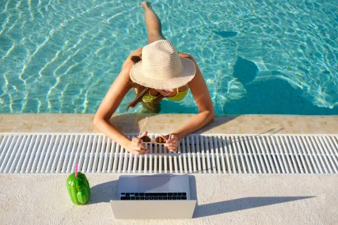 A woman is laying on the edge of a swimming pool using a laptop.
