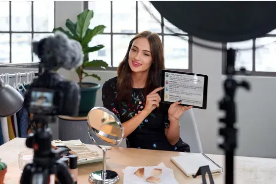 A woman is sitting at a table holding a tablet in front of a camera.