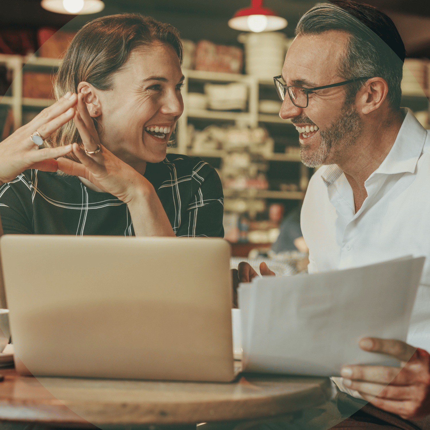 Two smiling coworkers discussing papers at a laptop in a café
