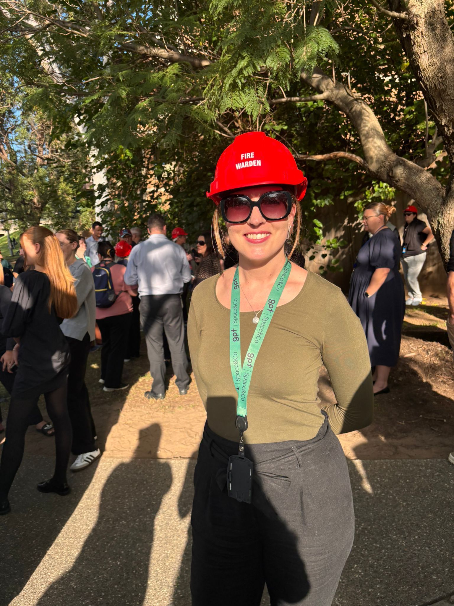Person in red hard hat and sunglasses standing outdoors at a crowded event in dappled sunlight
