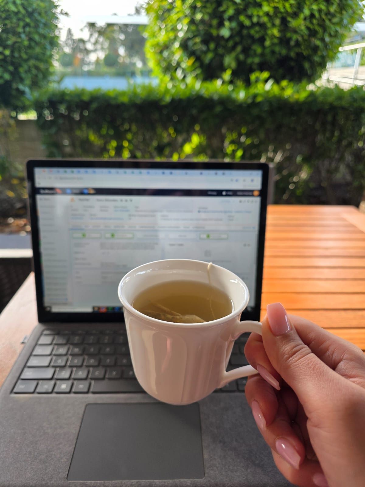 Hand holding a white cup of tea in front of a laptop on an outdoor wooden table