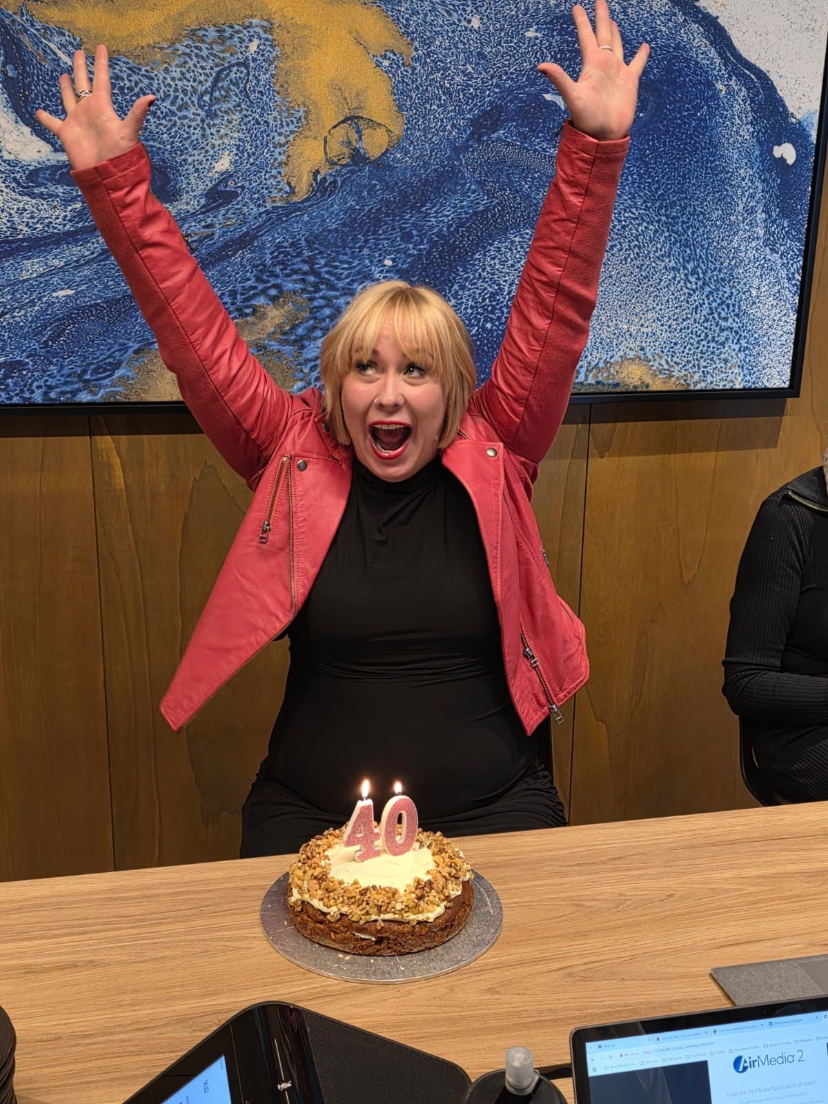Woman in a red jacket with arms raised, celebrating beside a lit birthday cake at a table
