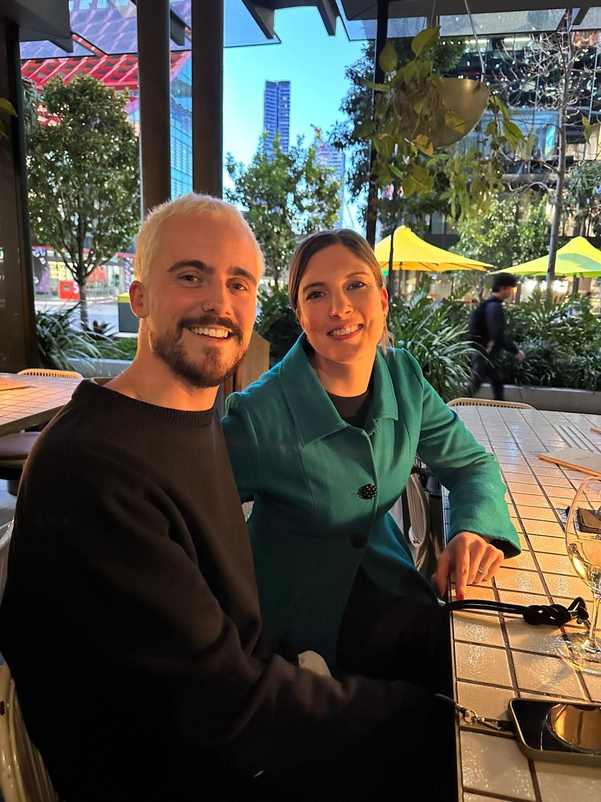 Two people seated at an outdoor café table, smiling in sunlight with plants and umbrellas behind them