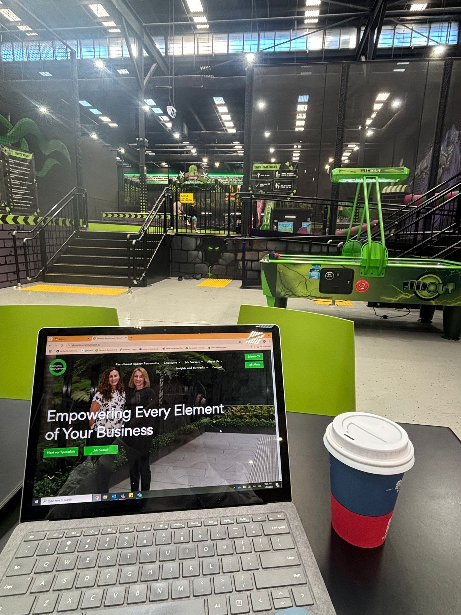 Laptop on a table in an industrial gym, with a coffee cup in front and green workout equipment in the background