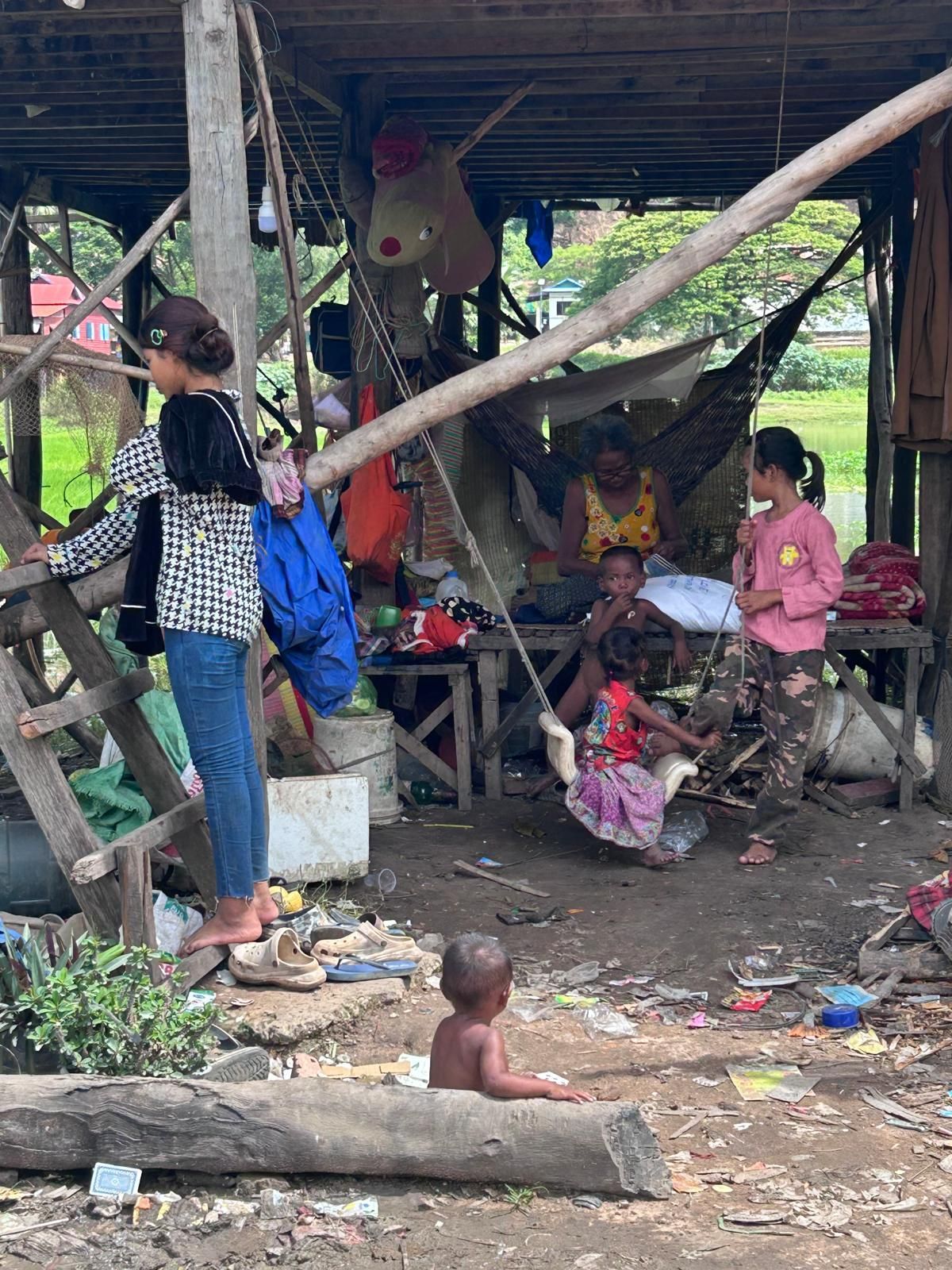 Children play in a cluttered, open-air shelter; one swings, others watch.
