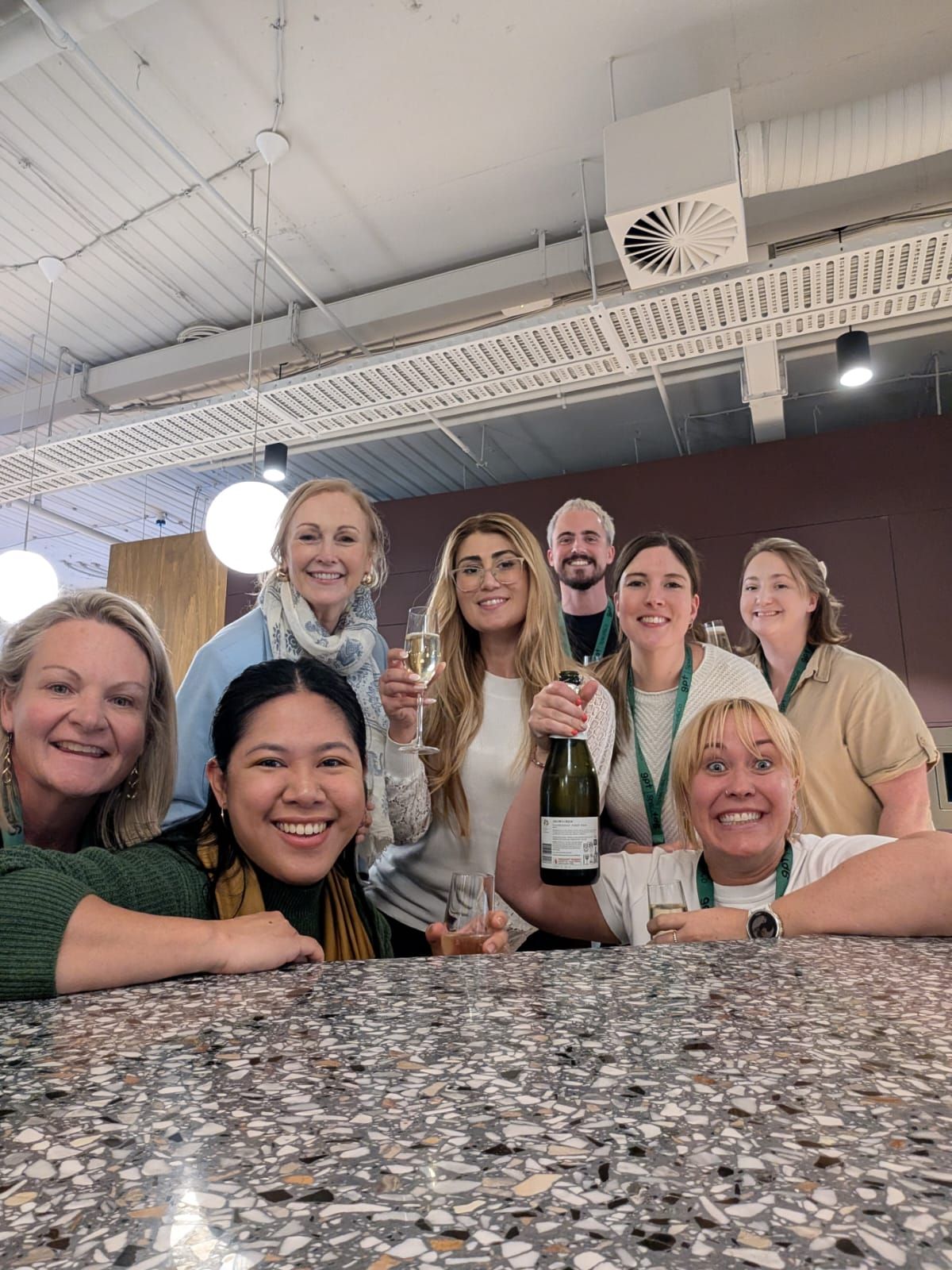 Group smiling at a restaurant table, one holding a champagne bottle under bright ceiling lights.