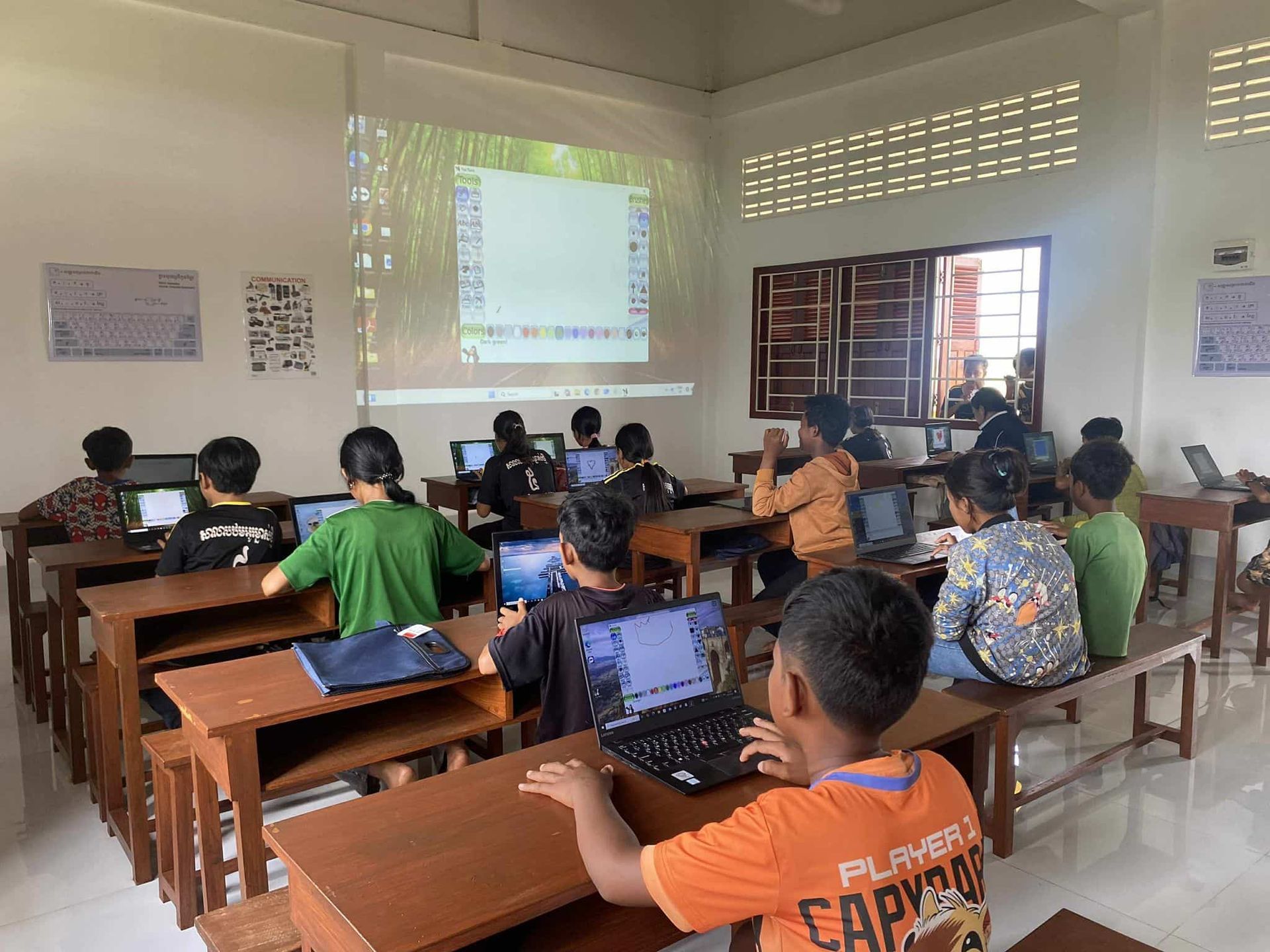 Classroom with students using laptops, a projector screen showing a desktop, and windows with natural light.