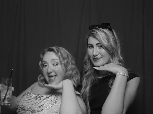 Two women posing playfully in front of a dark backdrop, one seated and one leaning with a hand under her chin.