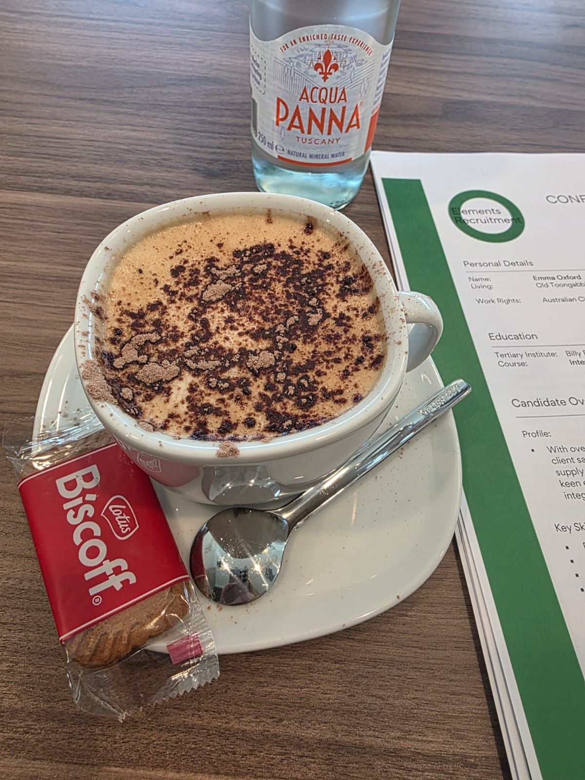Coffee cup with chocolate sprinkles on a saucer, spoon, Biscoff biscuit, and water bottle on a table