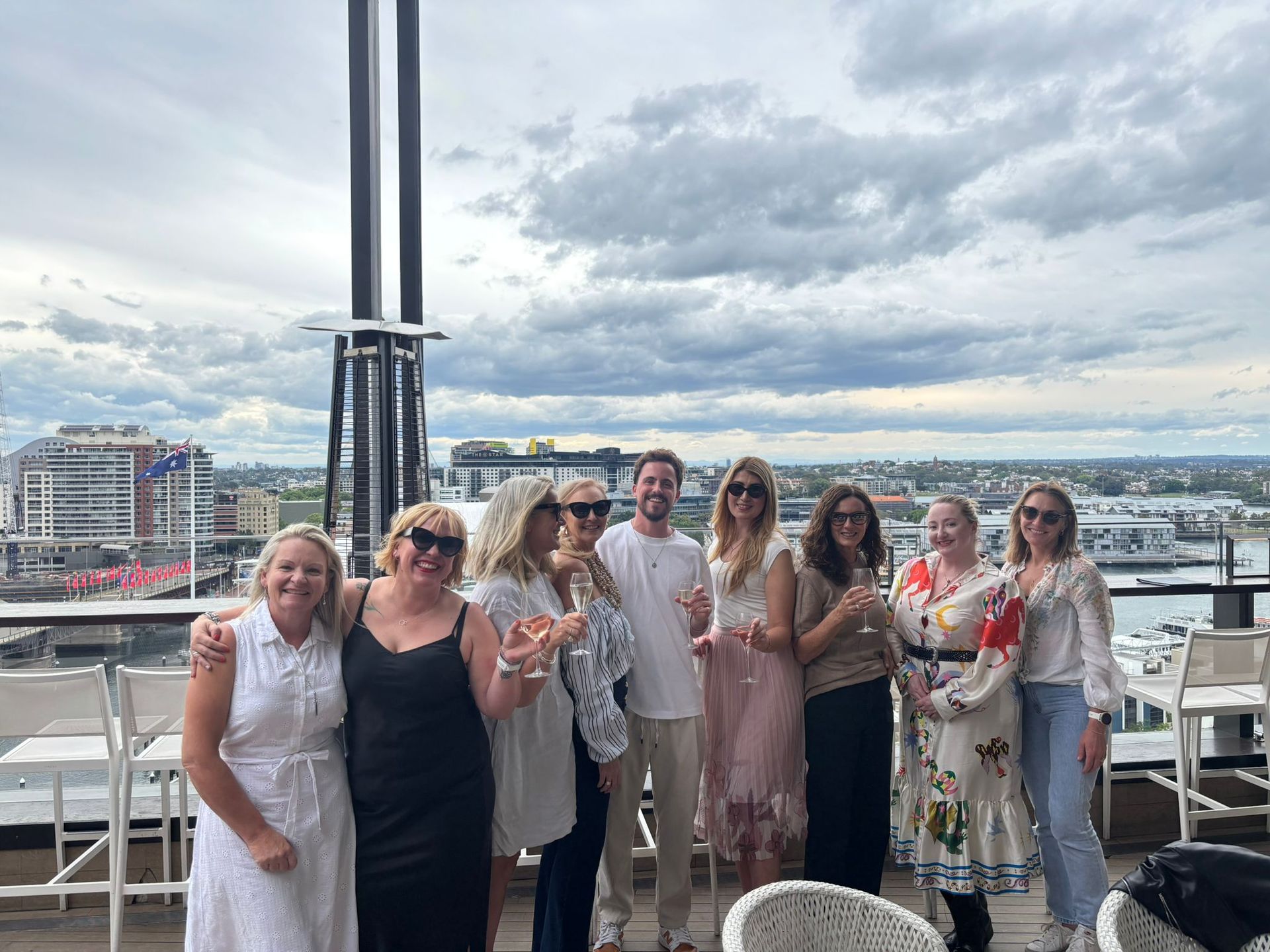Group of people posing on a rooftop terrace with a cloudy city skyline behind them.