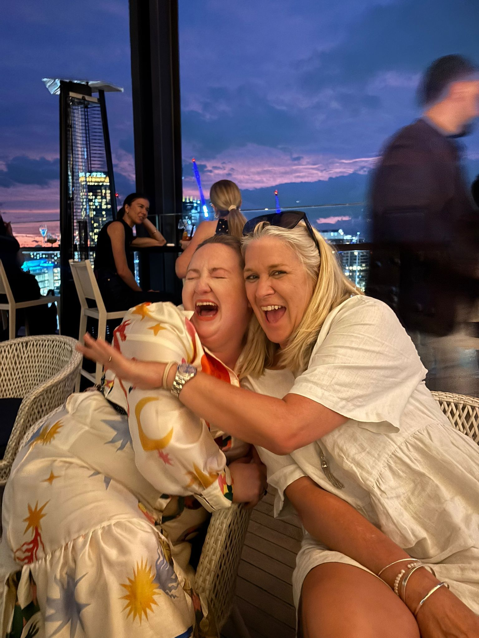 Two women laughing and hugging on a rooftop at sunset, with city lights in the background.