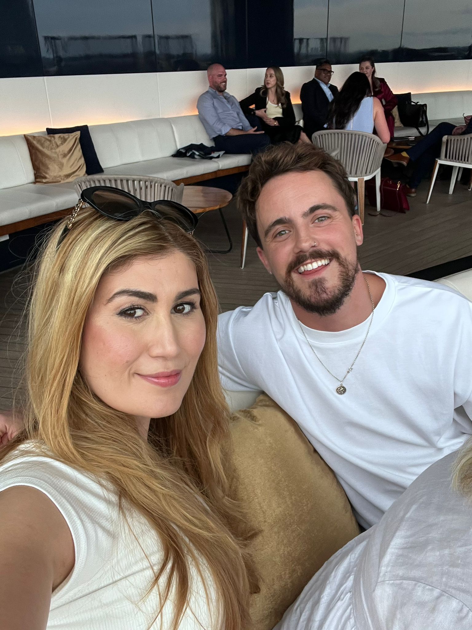 Two smiling people taking a selfie on a couch in a modern lounge, with seated guests in the background