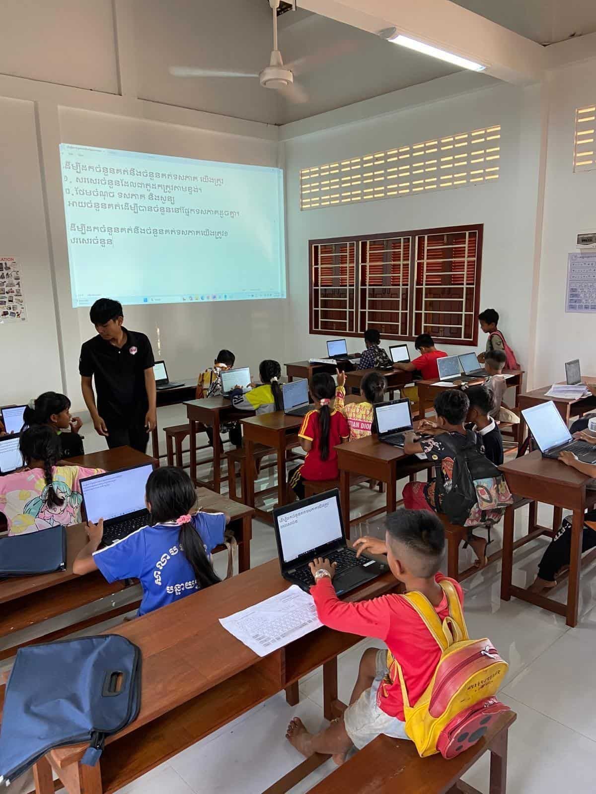 Classroom with students using laptops; teacher at front, lesson on screen, natural light.