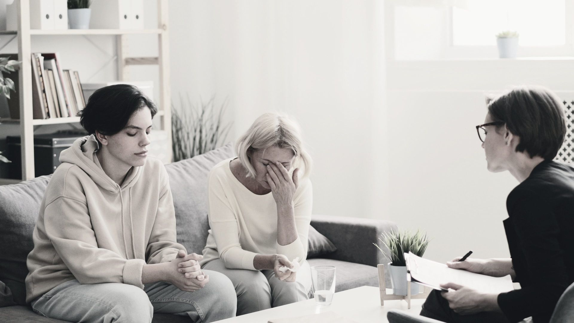 Three people in a bright room sit in a counseling session, one holding their head in distress.