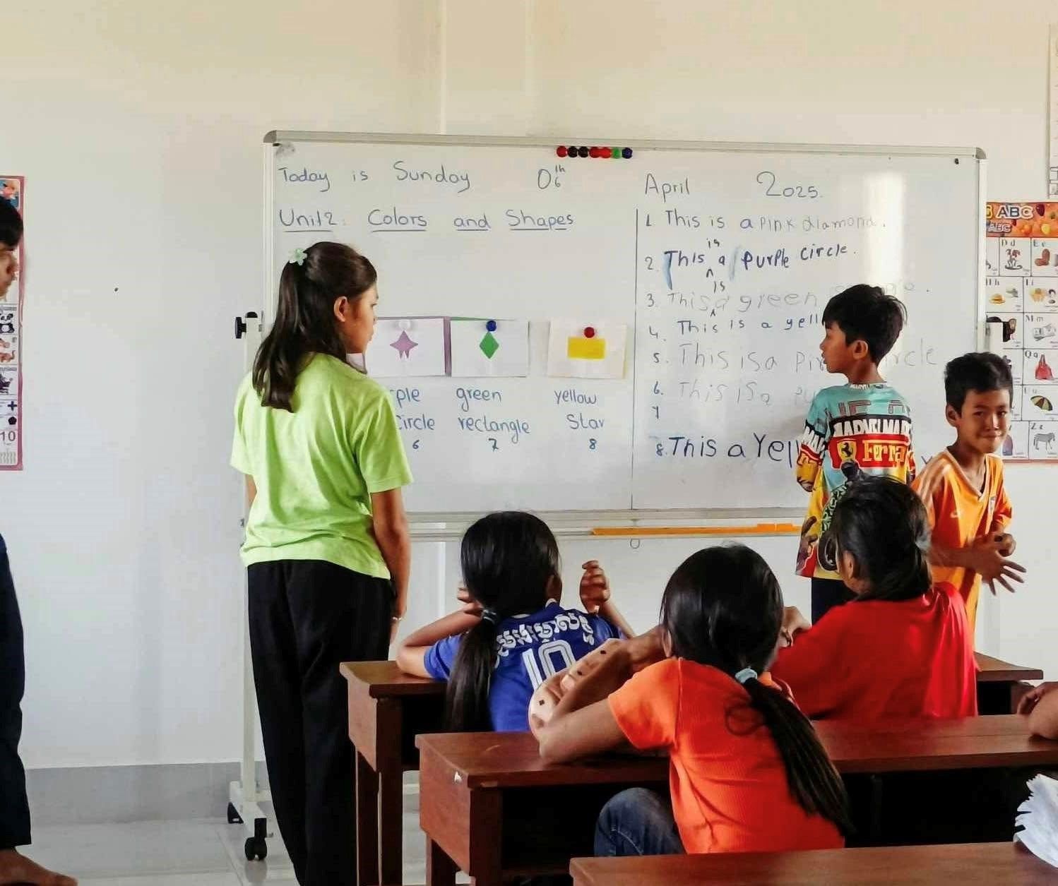 Teacher and students at a whiteboard in a classroom, with colorful learning materials on the wall.