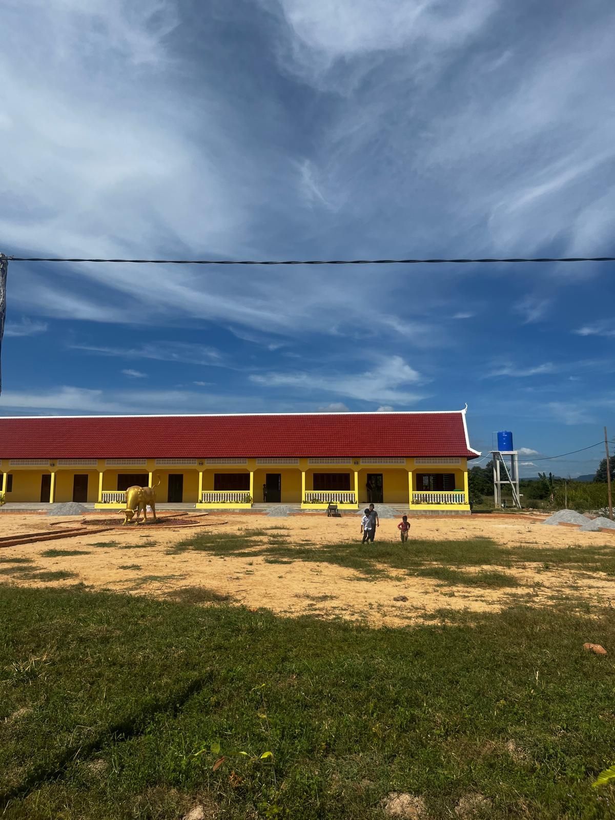 Yellow school building with red roof under a cloudy sky, viewed across a grassy dirt field