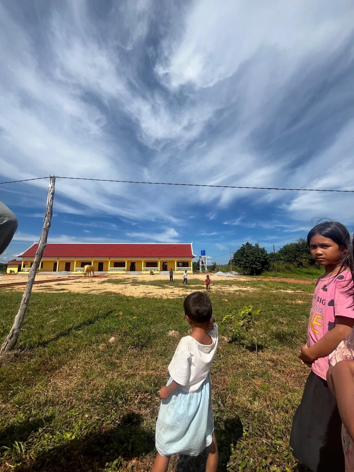 Two children stand in a grassy field near a red-roofed building under a dramatic cloudy sky.