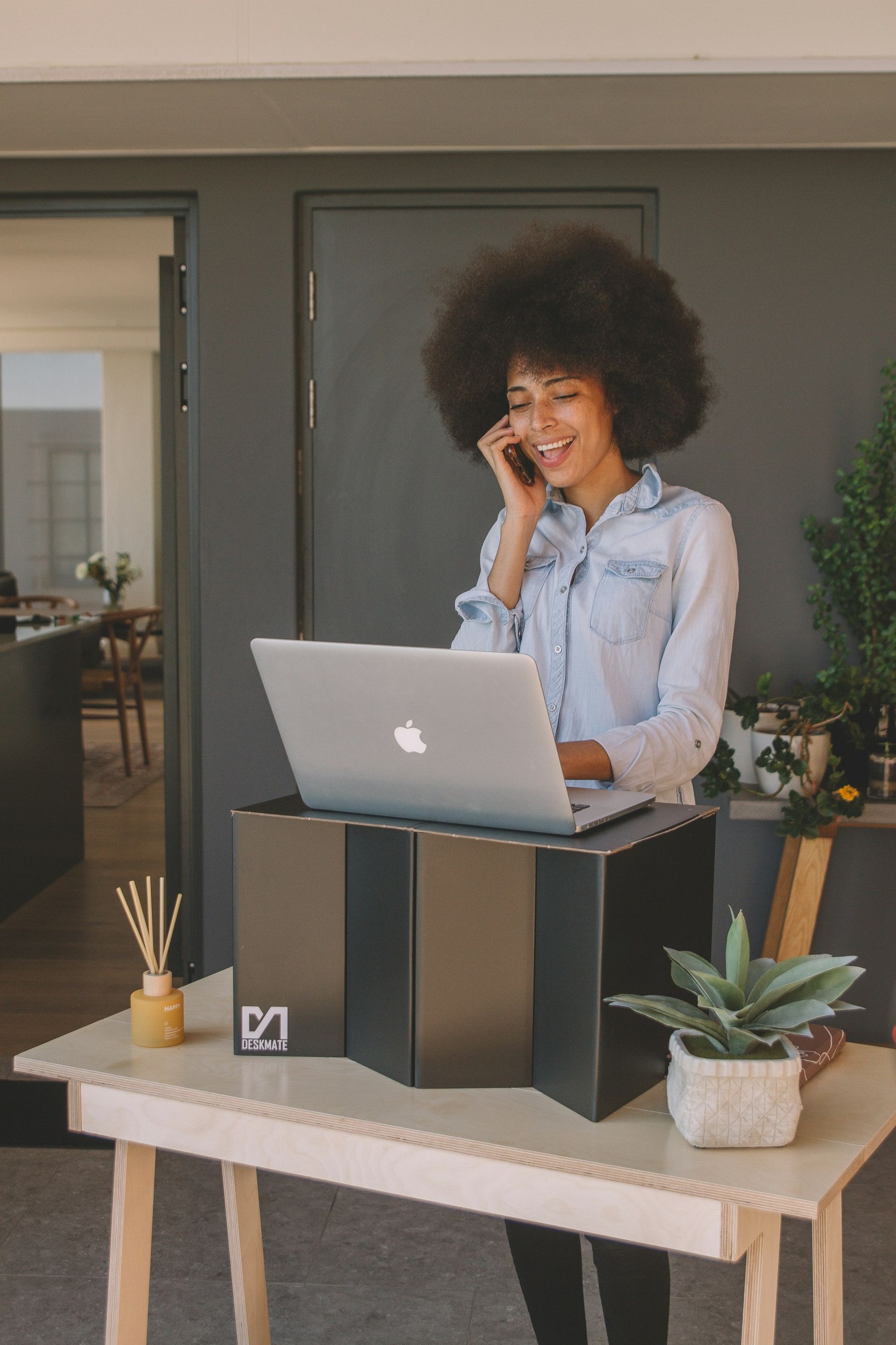 Person smiling on a phone call behind a laptop on a desk with plants indoors