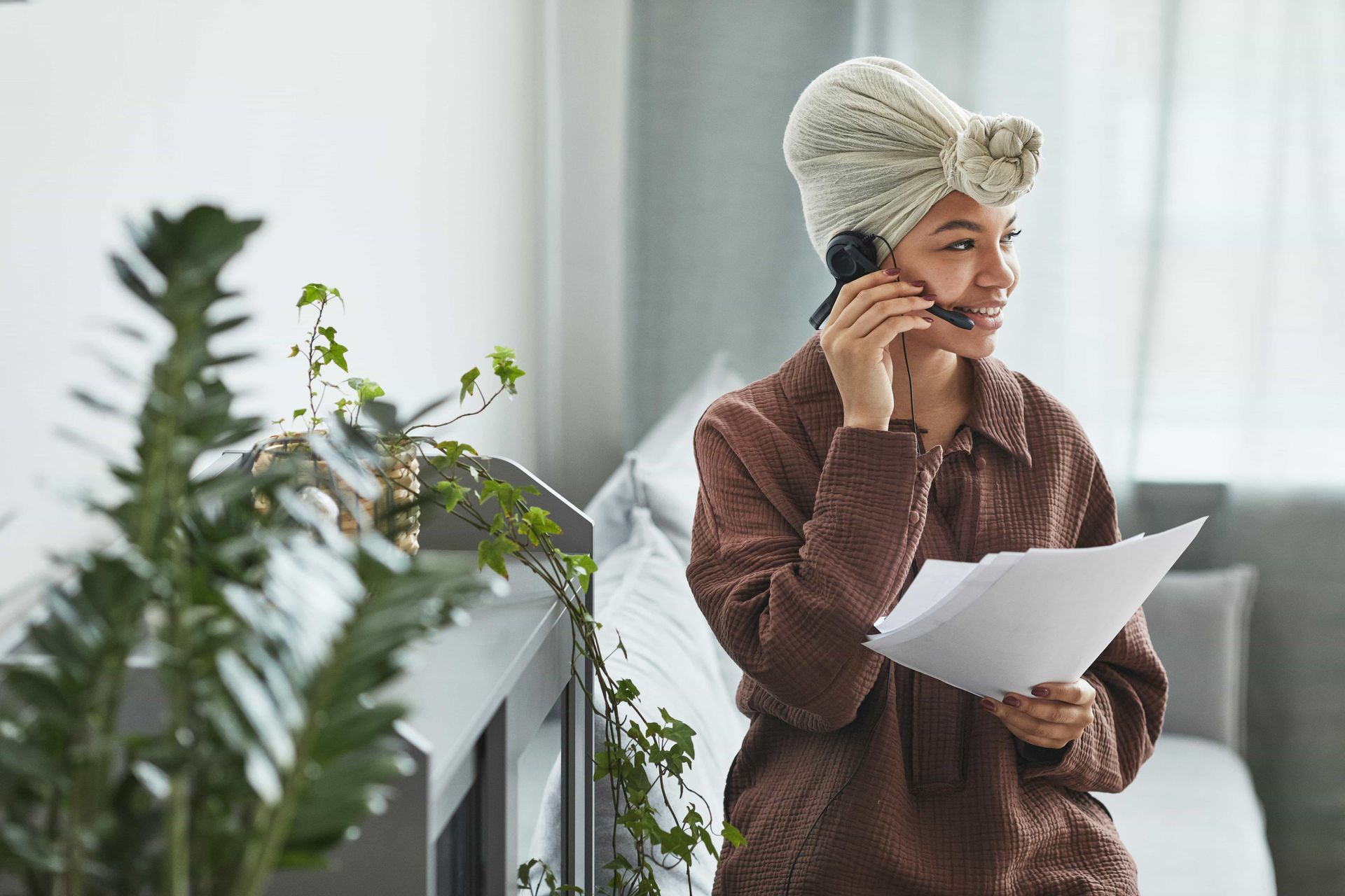Person in a brown robe and towel turban reads a paper while talking on the phone in a bright room.