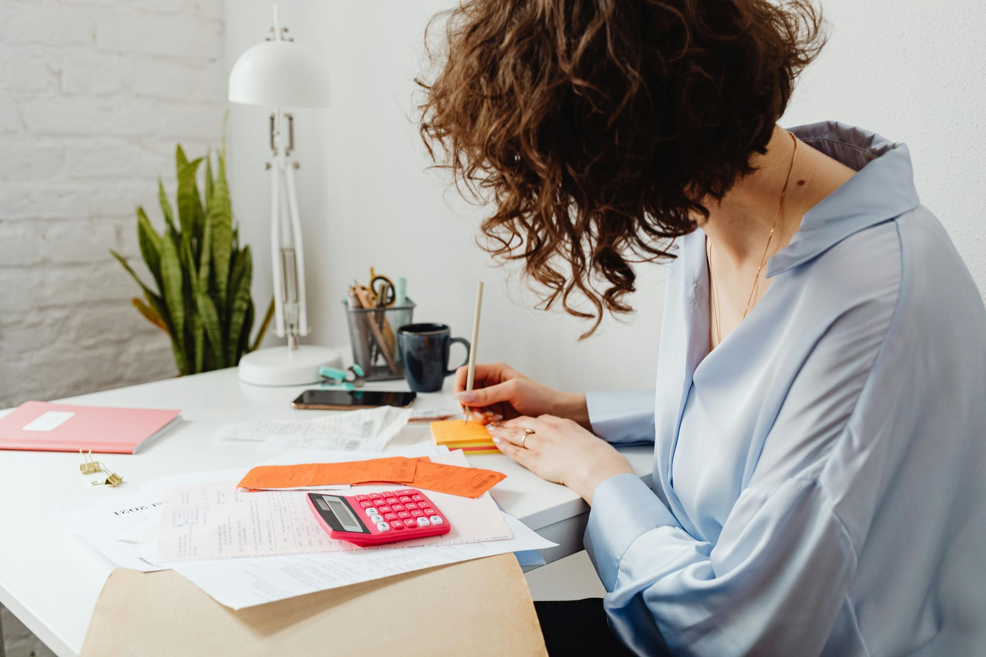 Person writing at a desk with papers, calculator, and office supplies in a bright workspace
