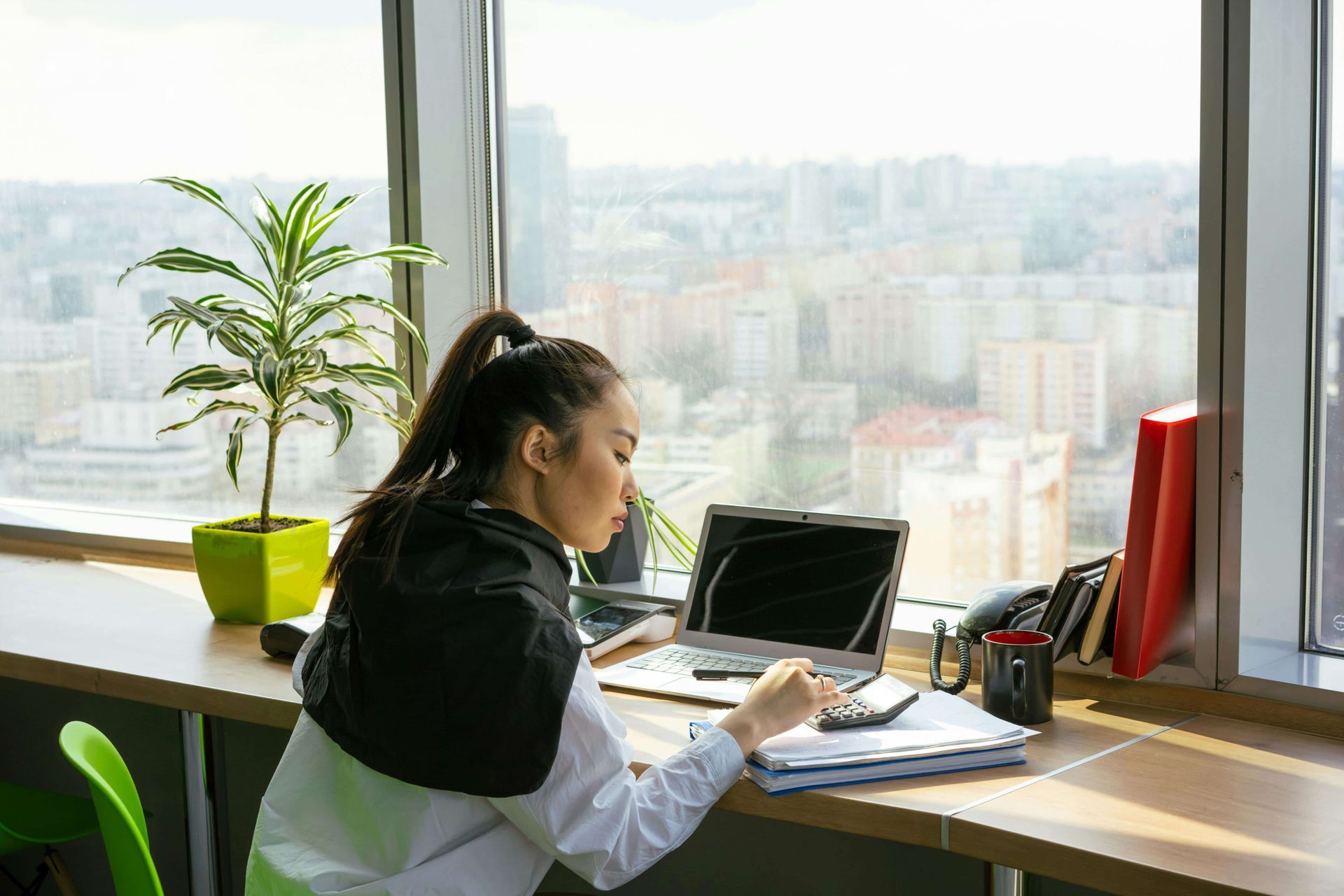 Person working at a desk by a bright window with a laptop, notebook, and potted plant