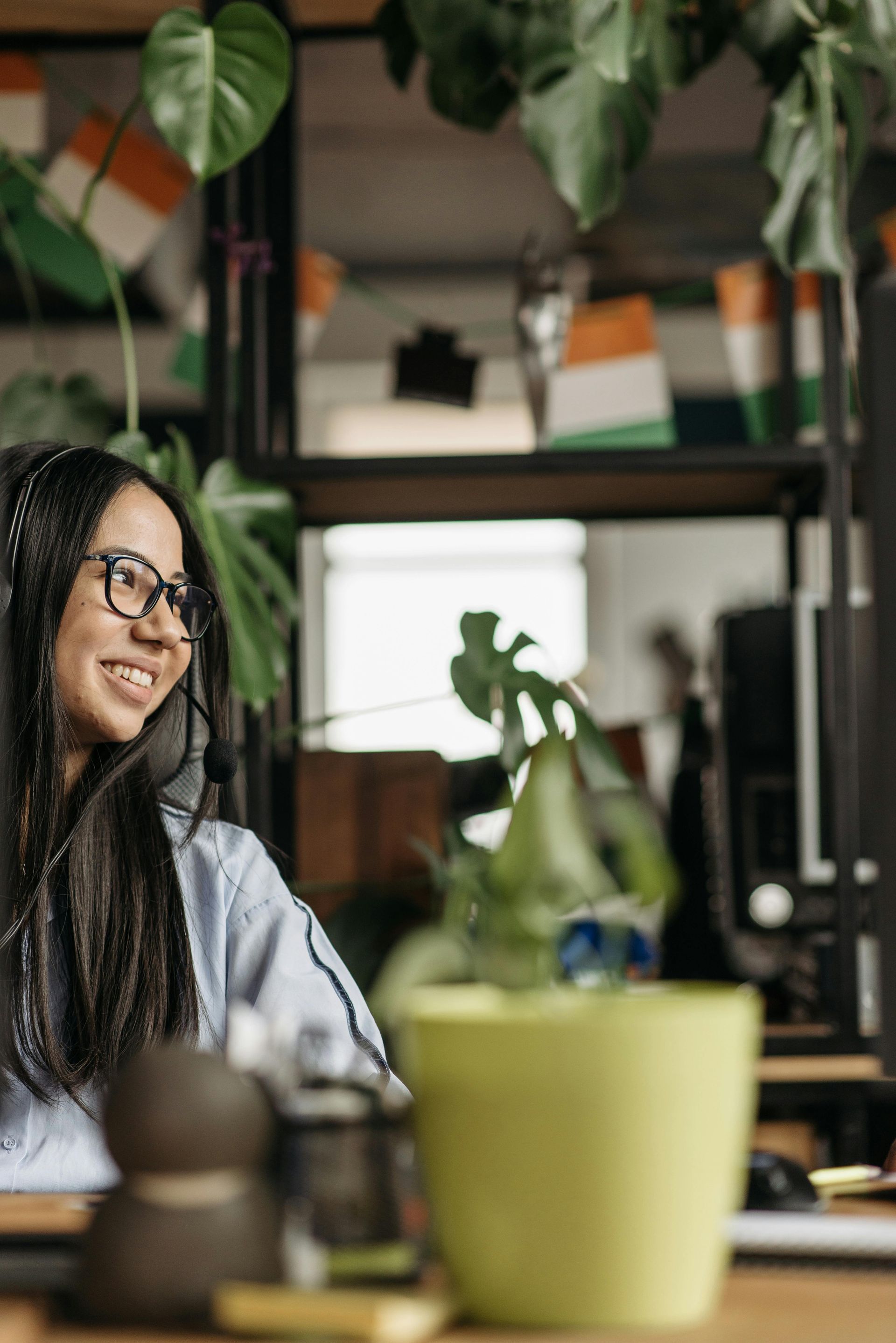 Woman smiling in an office with plants and shelves, seen beside a laptop and yellow mug