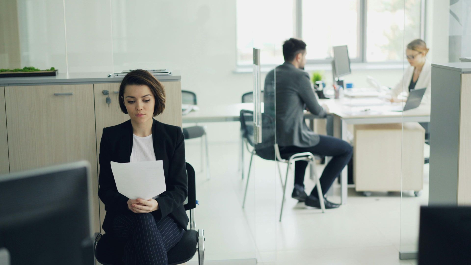 Woman in dark suit reading a paper in a bright office while two coworkers talk in the background