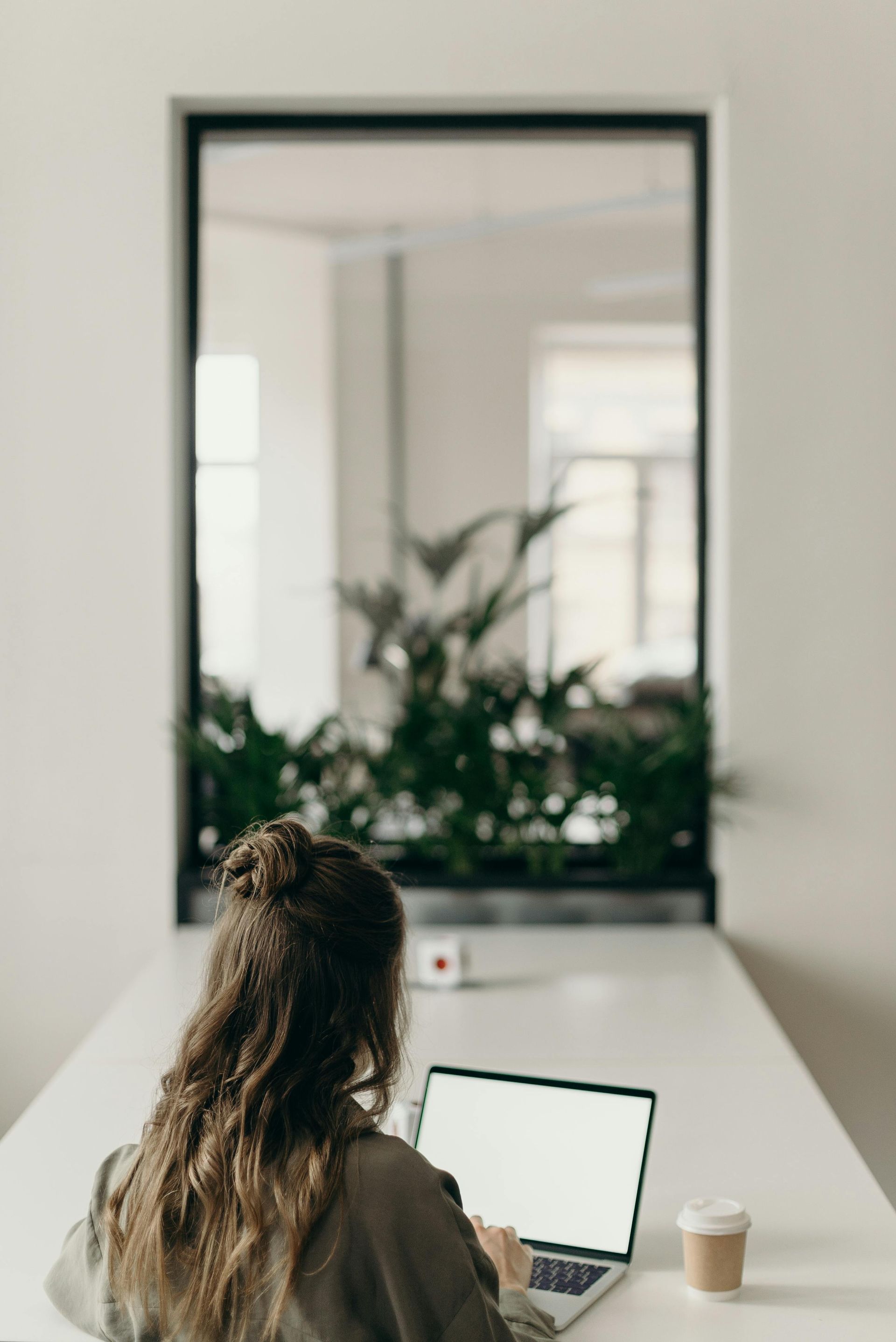 Person working on a laptop at a white desk in a bright room with a large mirror and plants