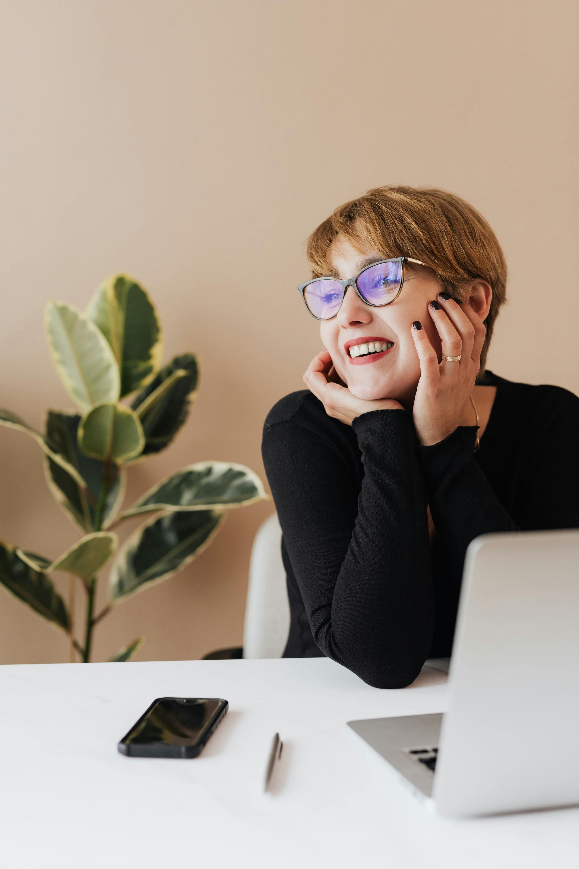 Person smiling at a desk with a laptop, phone, and plant, resting chin on hand