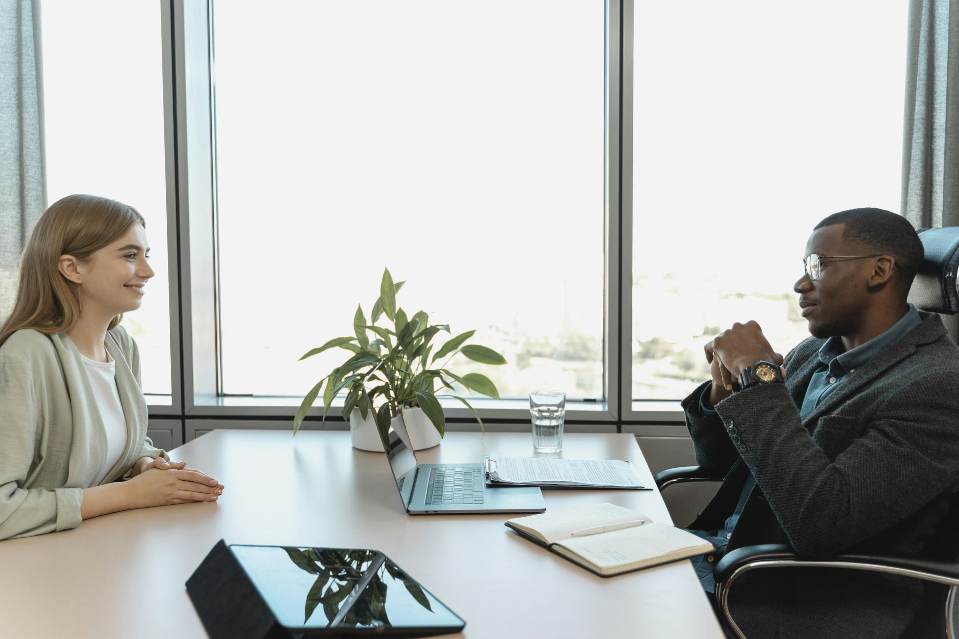 Two people in a bright office meeting across a table with a laptop, notebook, and plant.