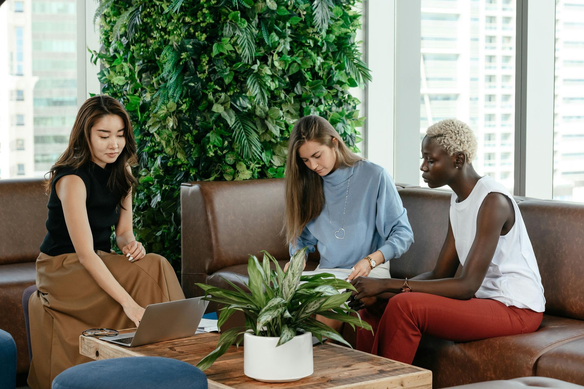 Three people collaborating with a laptop in a modern lounge beside a leafy wall and coffee table