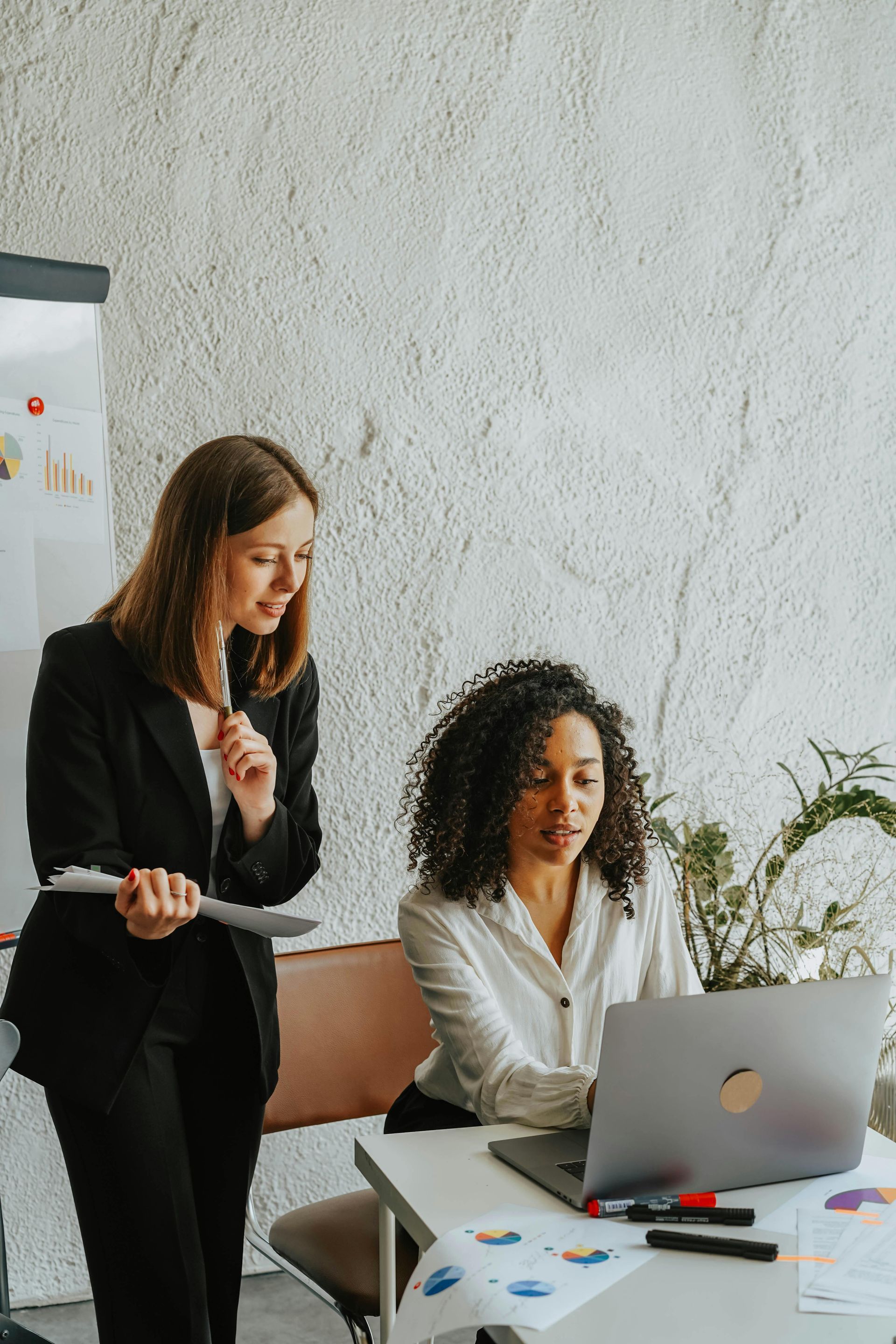 Two women in business attire at a desk, one looking at a laptop, the other looking on, holding a document.