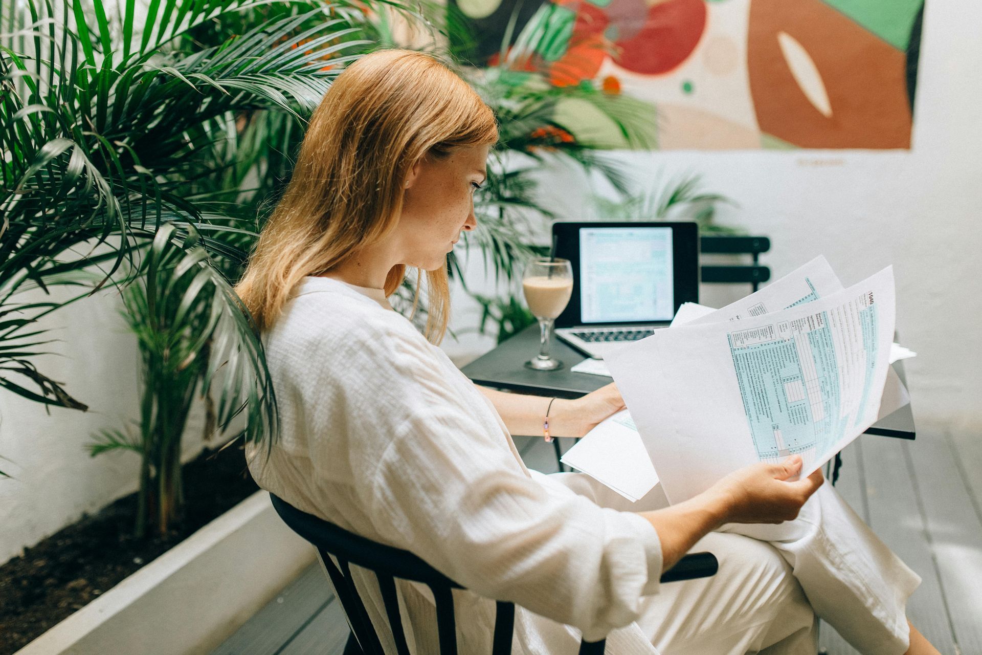 Person reading papers beside a laptop in a bright indoor workspace with plants