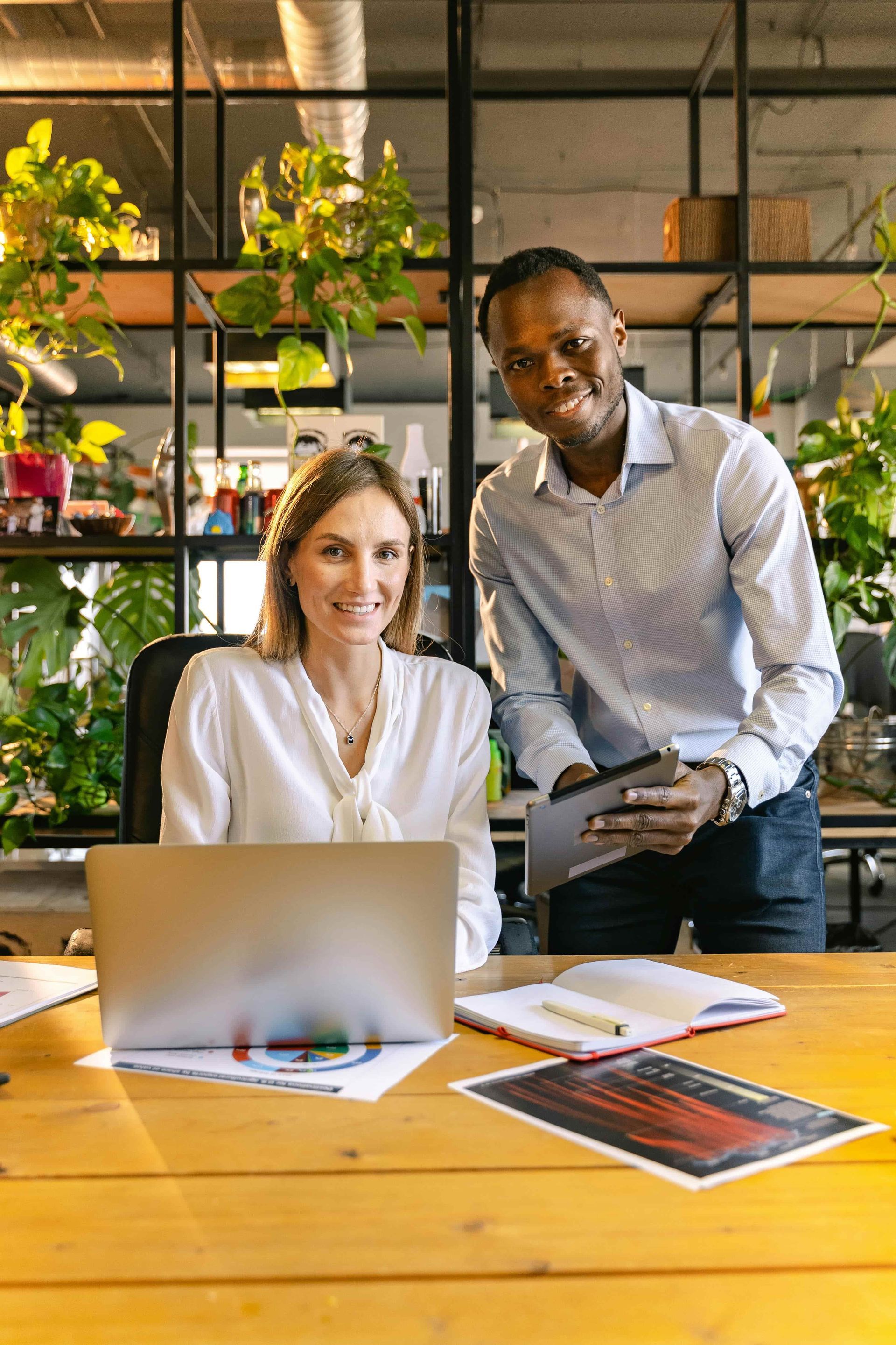 Two coworkers at a table with a laptop and papers in a plant-filled office