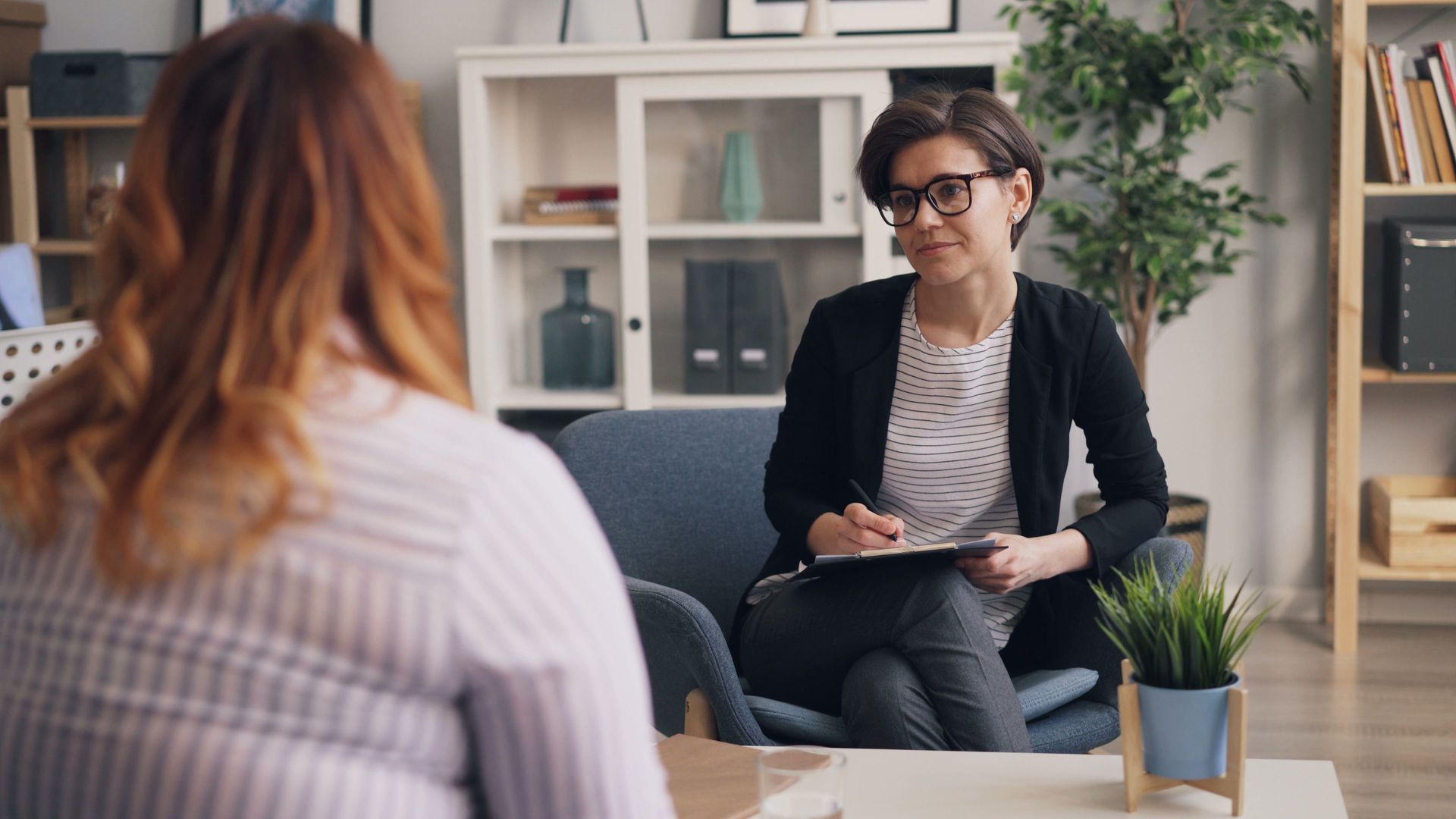 Two people in a bright office during a seated session, one taking notes across from another.