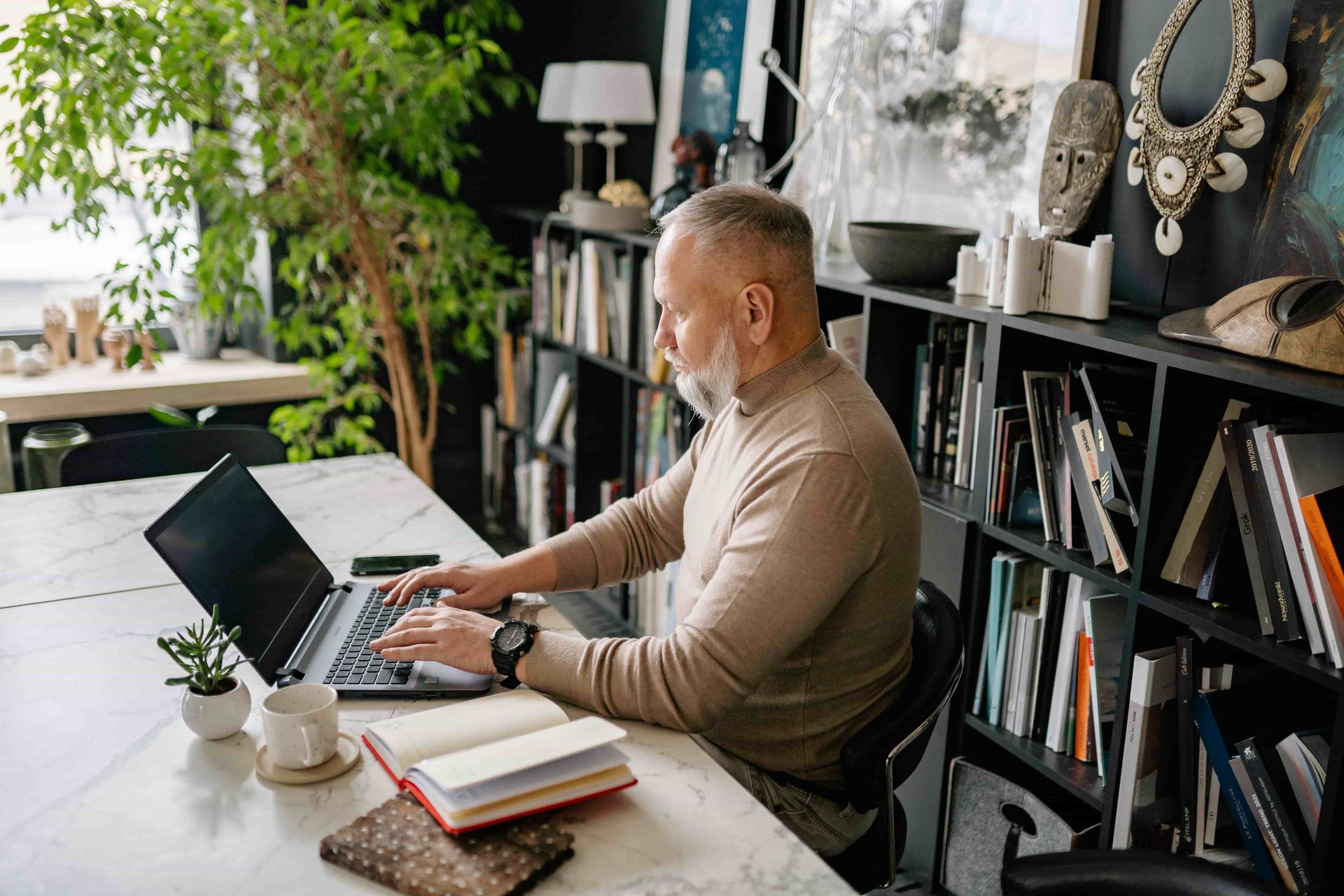 Person working on a laptop at a desk in a cozy home office with bookshelves and plants