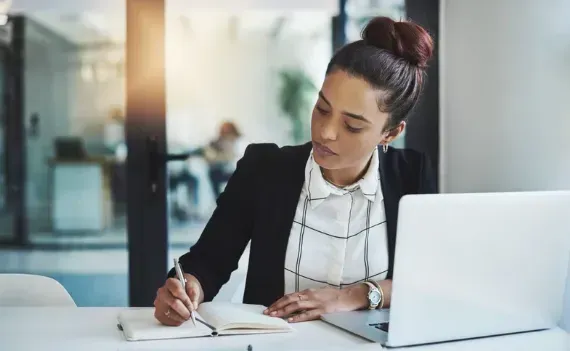 A woman is sitting at a desk with a laptop and writing in a notebook.