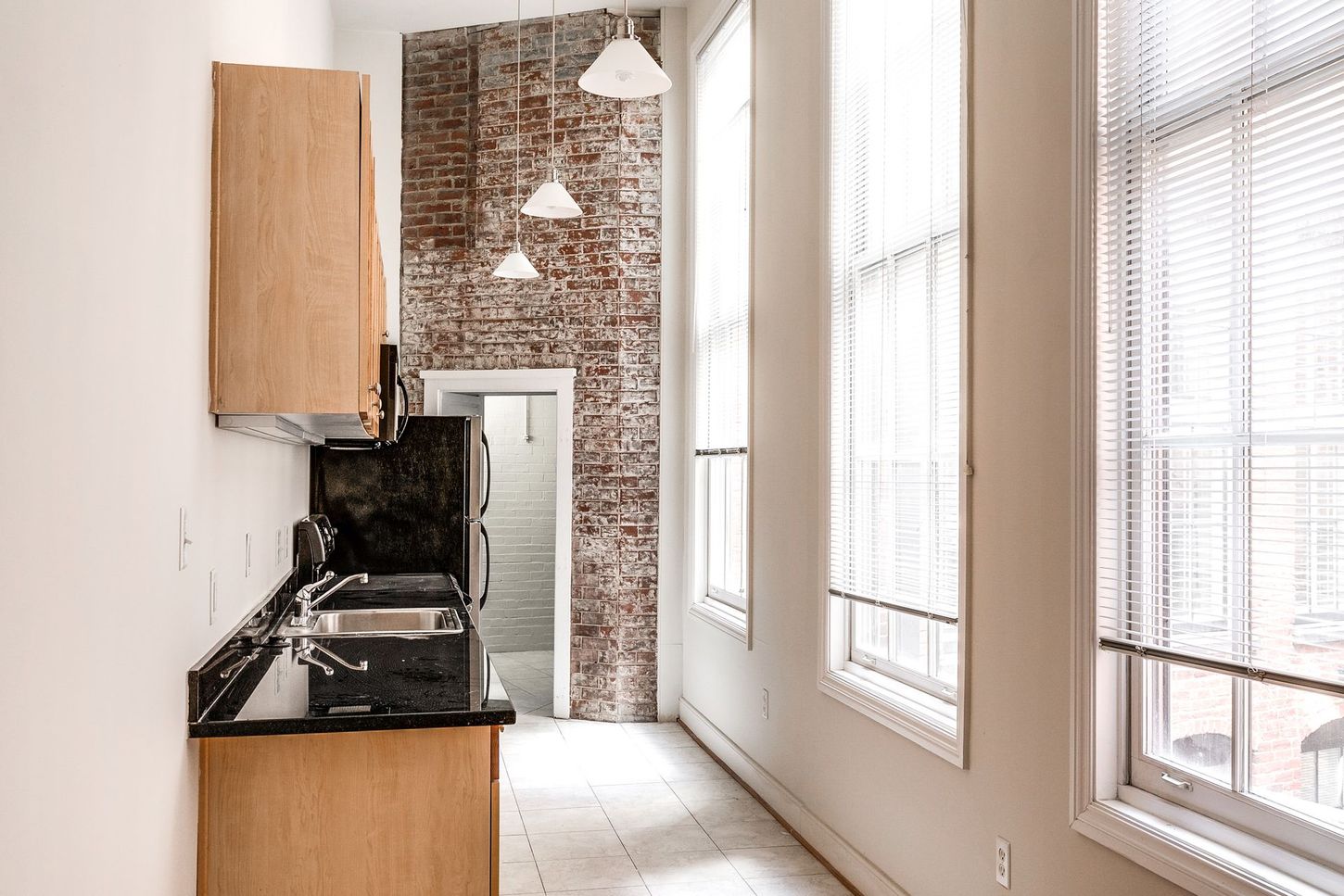 Kitchen with exposed brick wall, three hanging lights, and tall windows with blinds.
