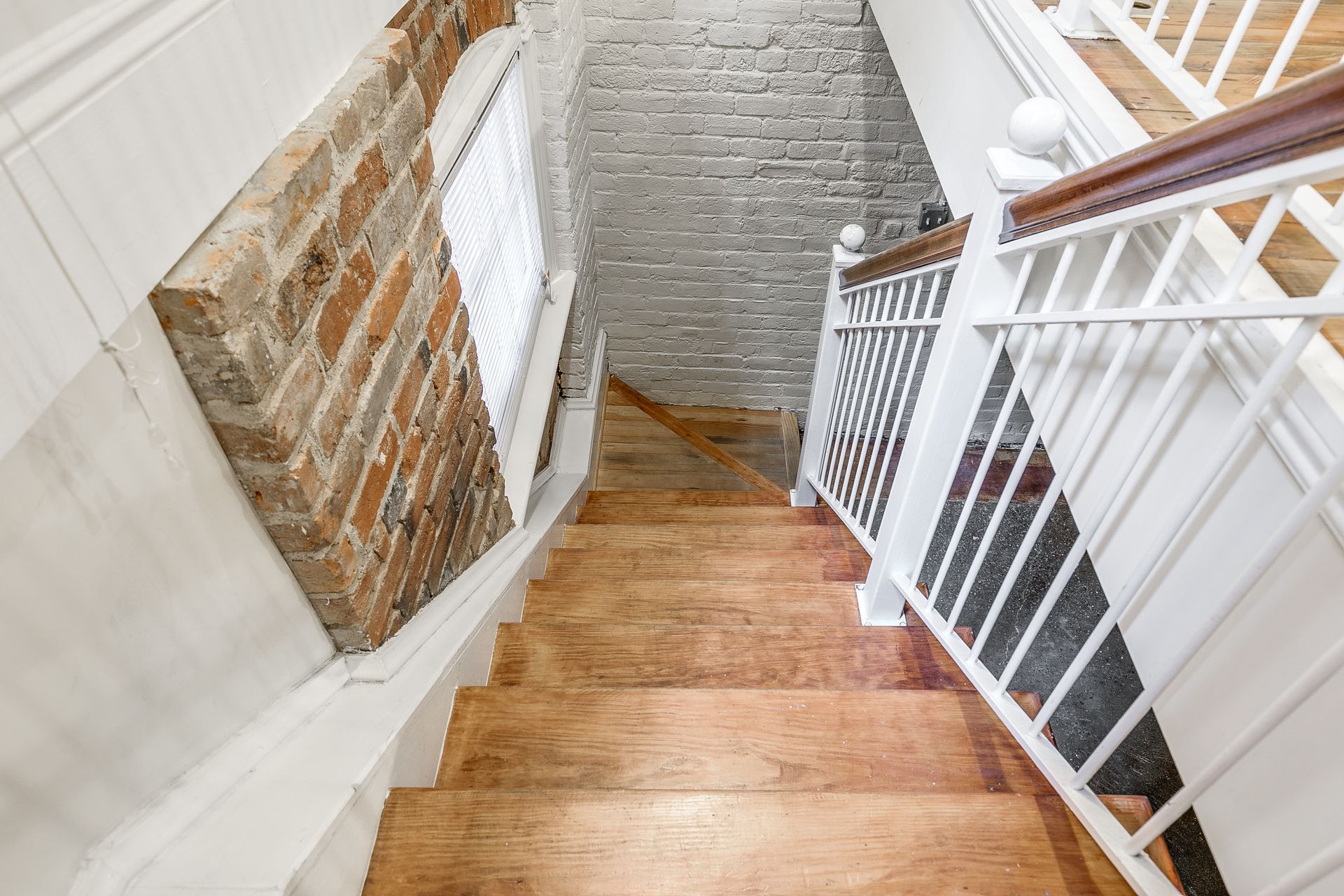 Wooden staircase descends between white railings, exposed brick wall, and natural light.