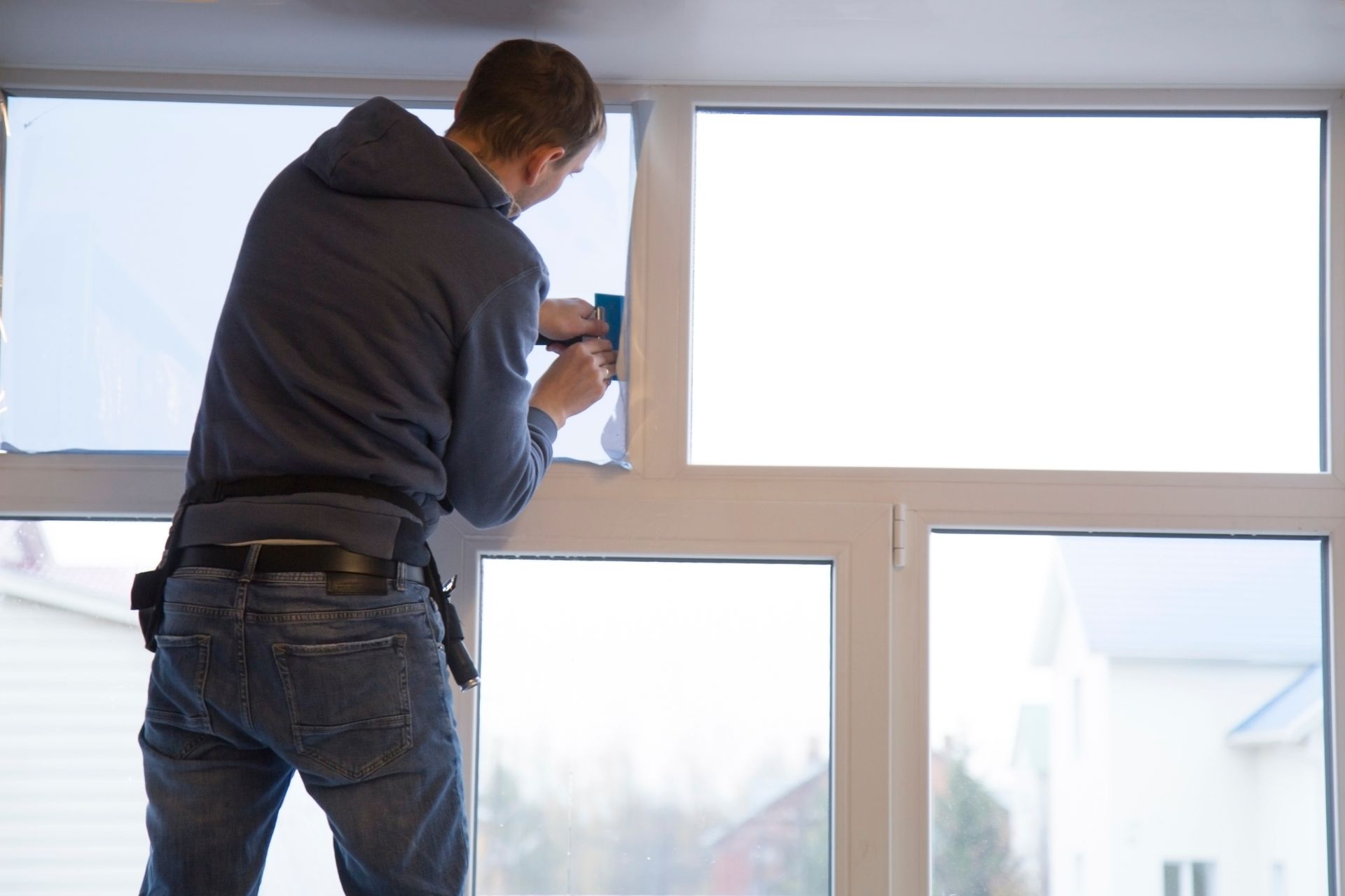 Installer in a hoodie applying protective film to a large white-framed window with a squeegee.