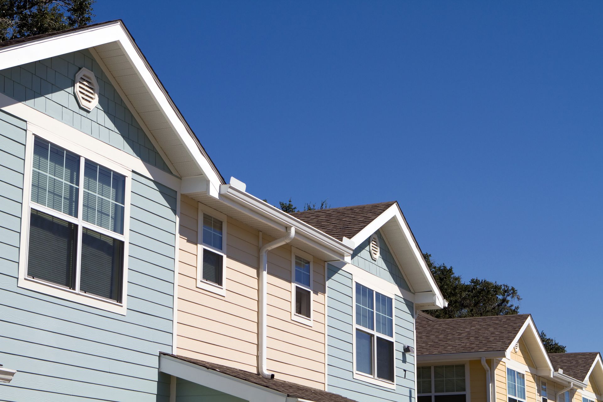 Side picture of an apartment with small windows with white frames, with the blue sky as background. Row of colorful houses with blue siding and brown roofs under a clear blue sky.
