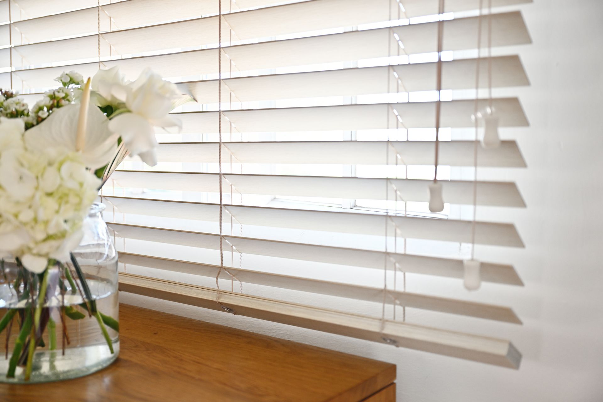 Wooden blinds next to a bouquet of white flowers in a glass vase, on a wooden table.