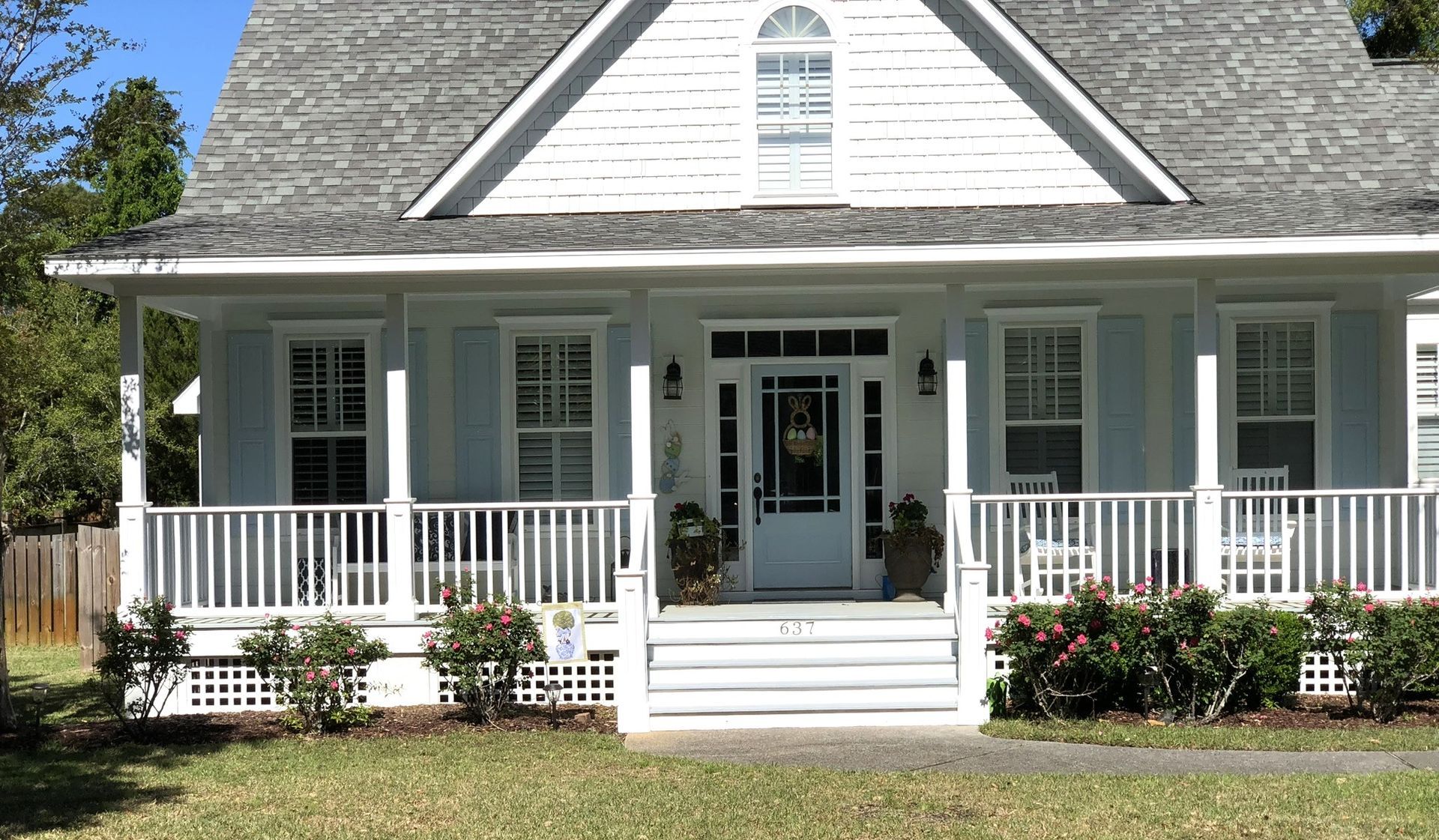 White house with a porch, blue shutters, gray roof, and green grass.