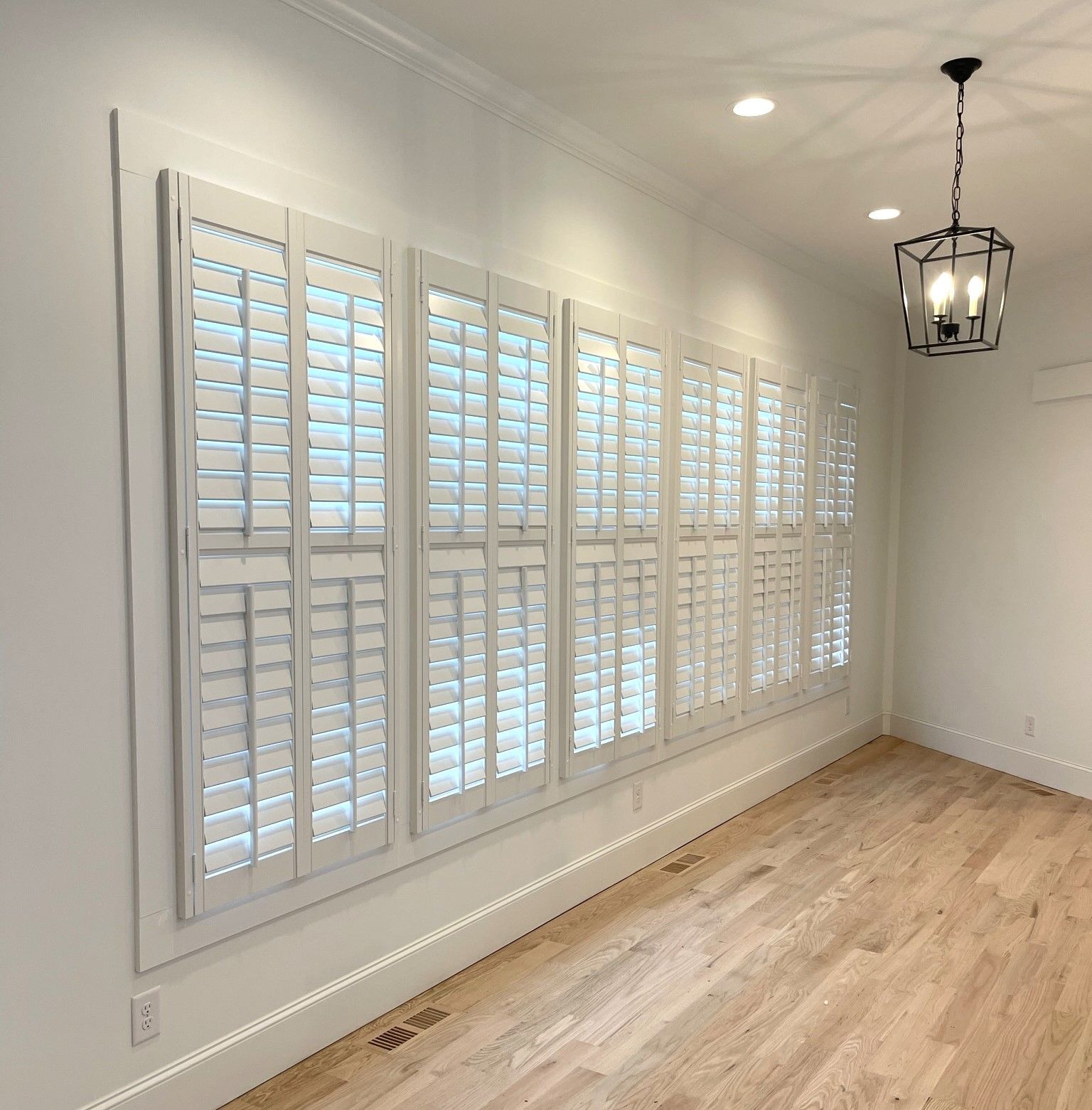 White shutters on a wall with light wood floors and a hanging black chandelier.